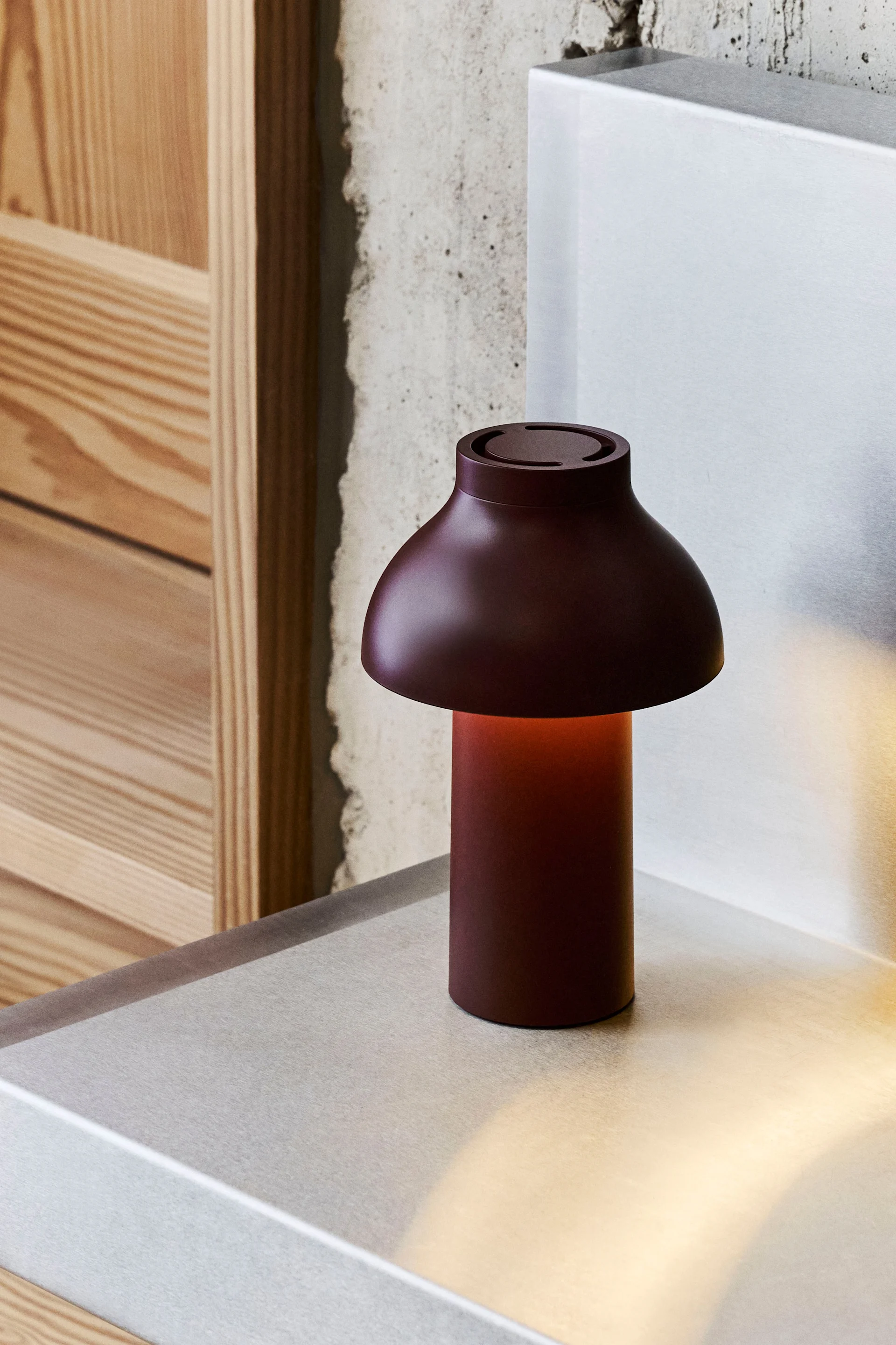 Een donkerrode paddestoelvormige tafellamp met een warme gloed op een metalen oppervlak, met beton en hout op de achtergrond.