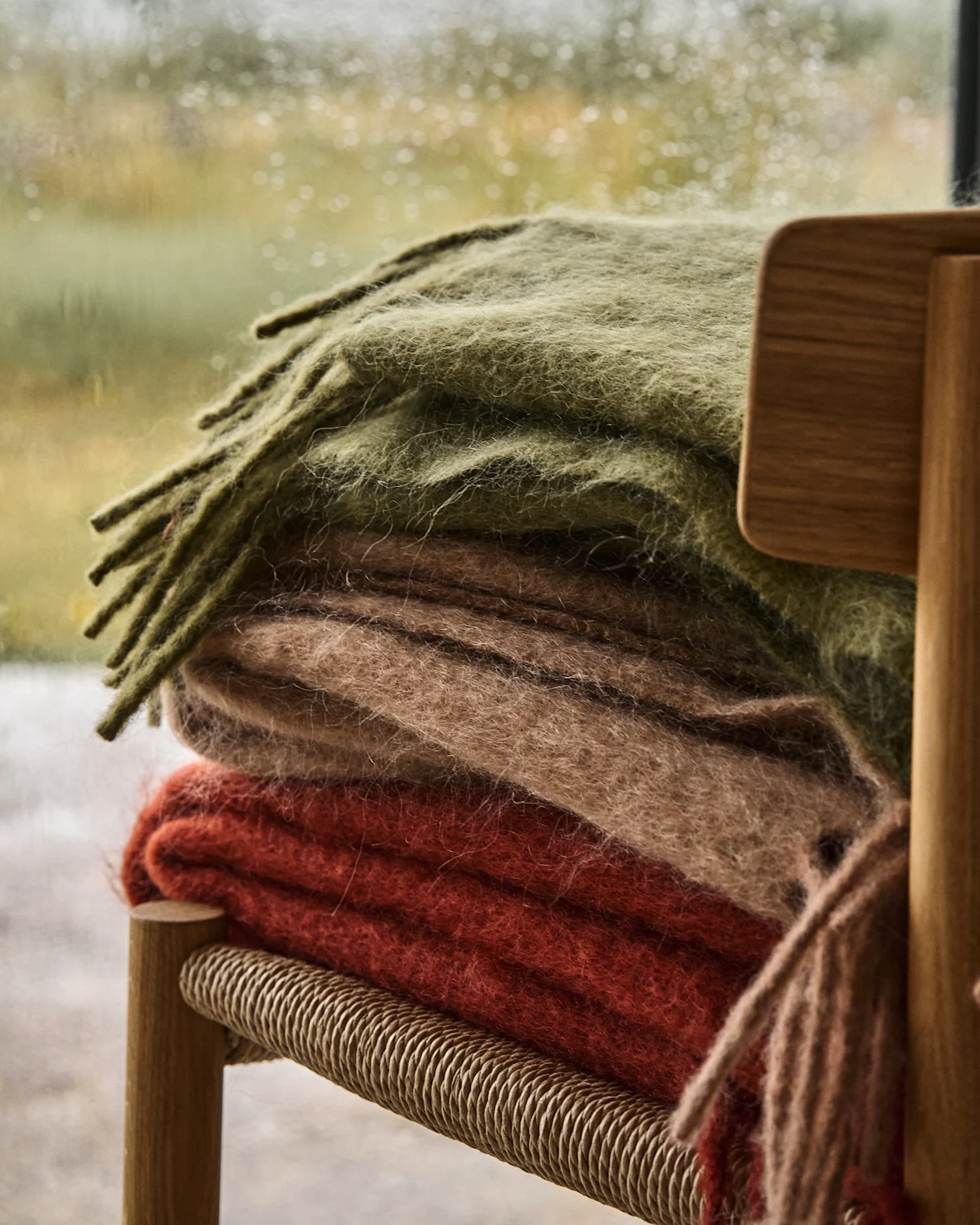 Stapel van drie zachte dekens in olijfgroen, beige en roodoranje op een houten stoel, met een regenachtig raam op de achtergrond.