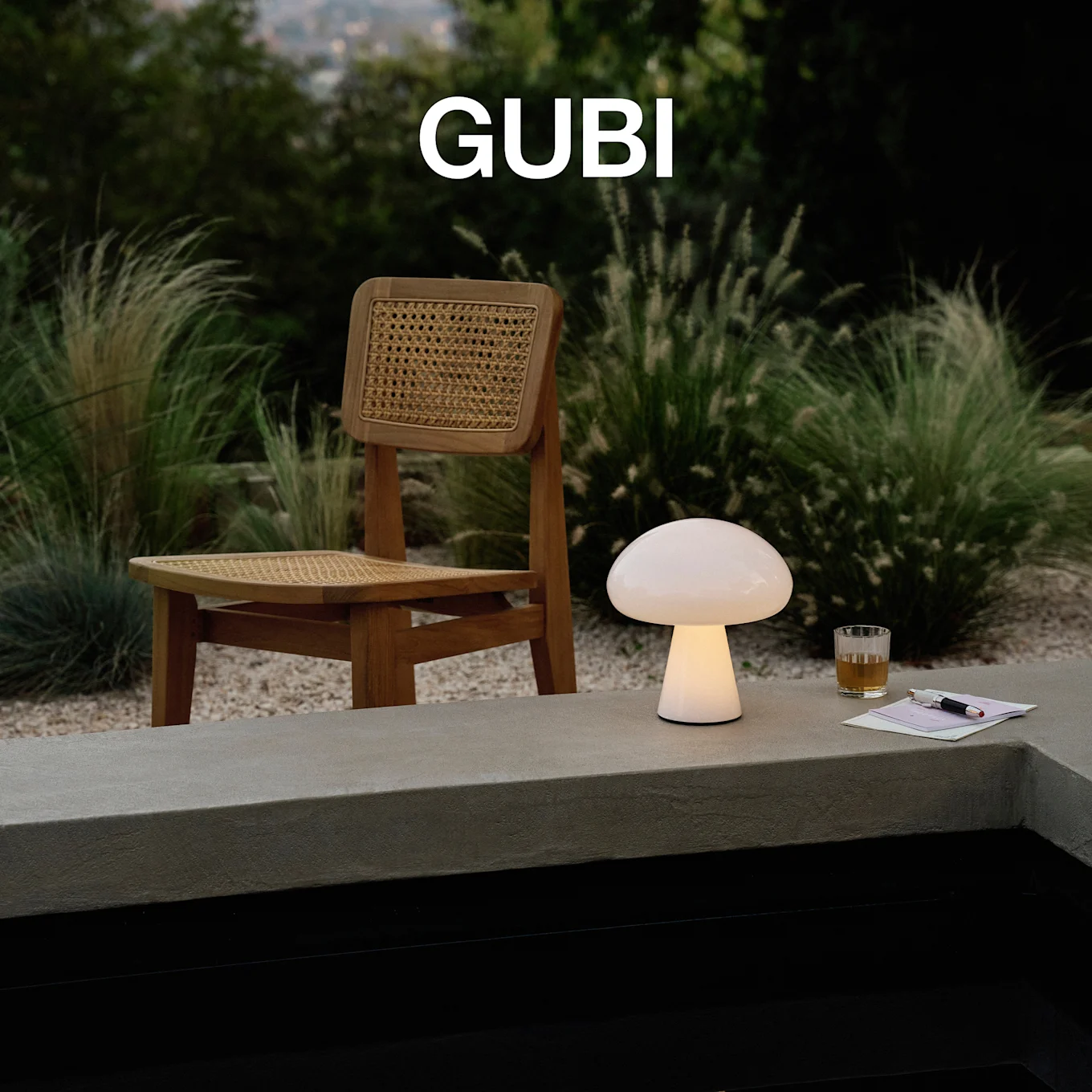 GUBI-logo. Een verlichte paddenstoelvormige tafellamp, een glas en een pen op een betonnen rand, met een houten stoel en weelderig groen.