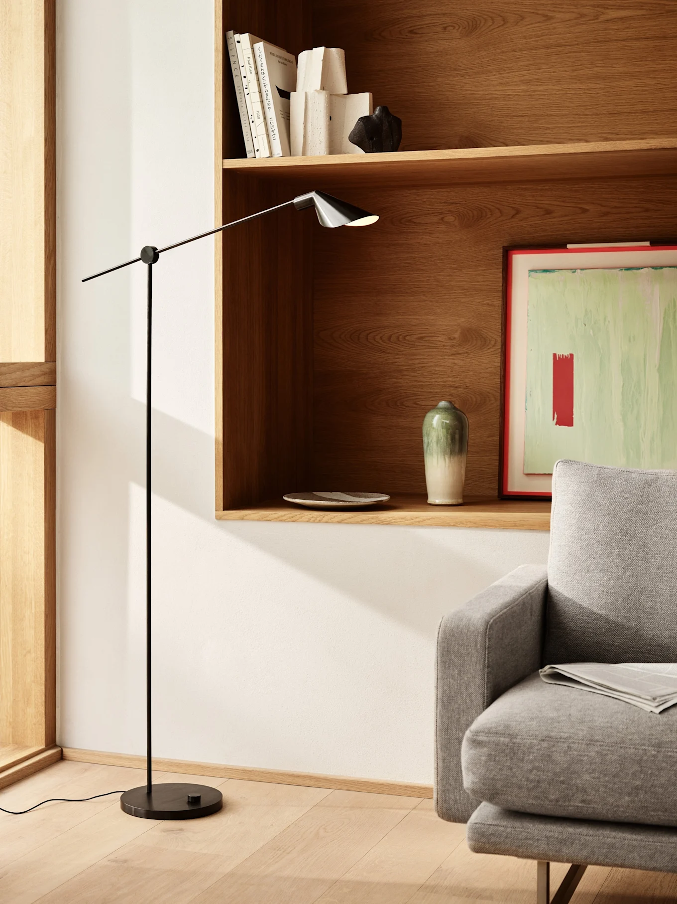 Een moderne woonkamer met een slanke zwarte vloerlamp, houten planken met boeken en decoratie, en een deel van een grijze bank.