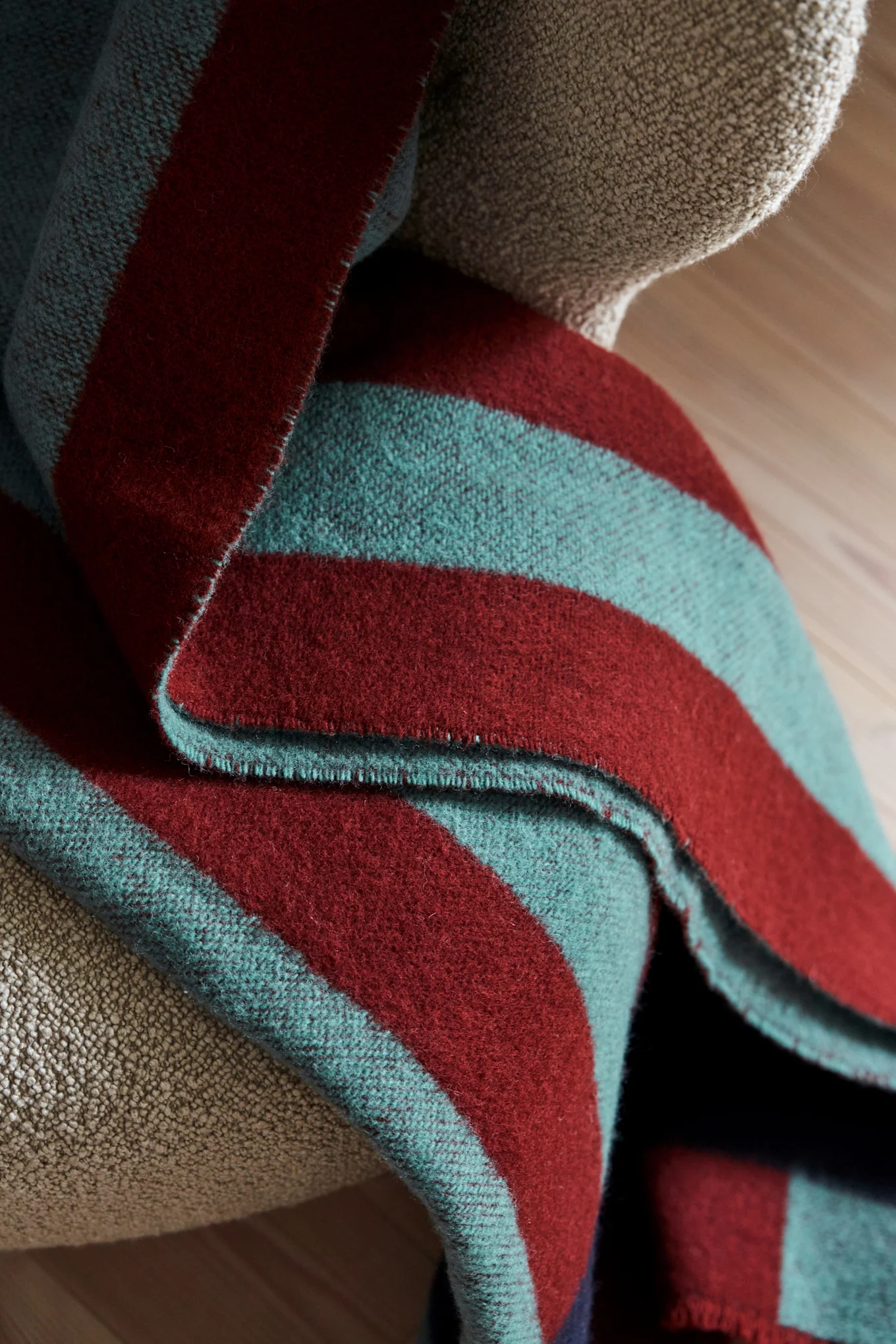 Gevouwen gestreepte deken met donkerrode en groenblauwe strepen gedrapeerd over een gestructureerde lichtbruine fauteuil.
