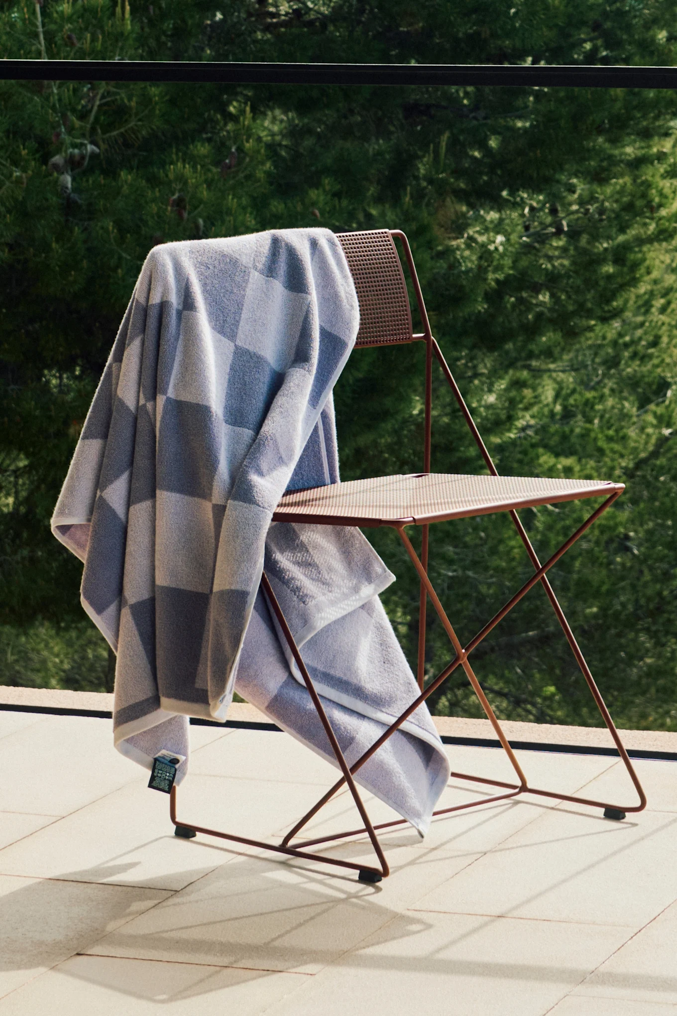 Lichtblauw en grijs geruite handdoek op een roodbruine metalen klapstoel op een zonnig terras met groen gebladerte.