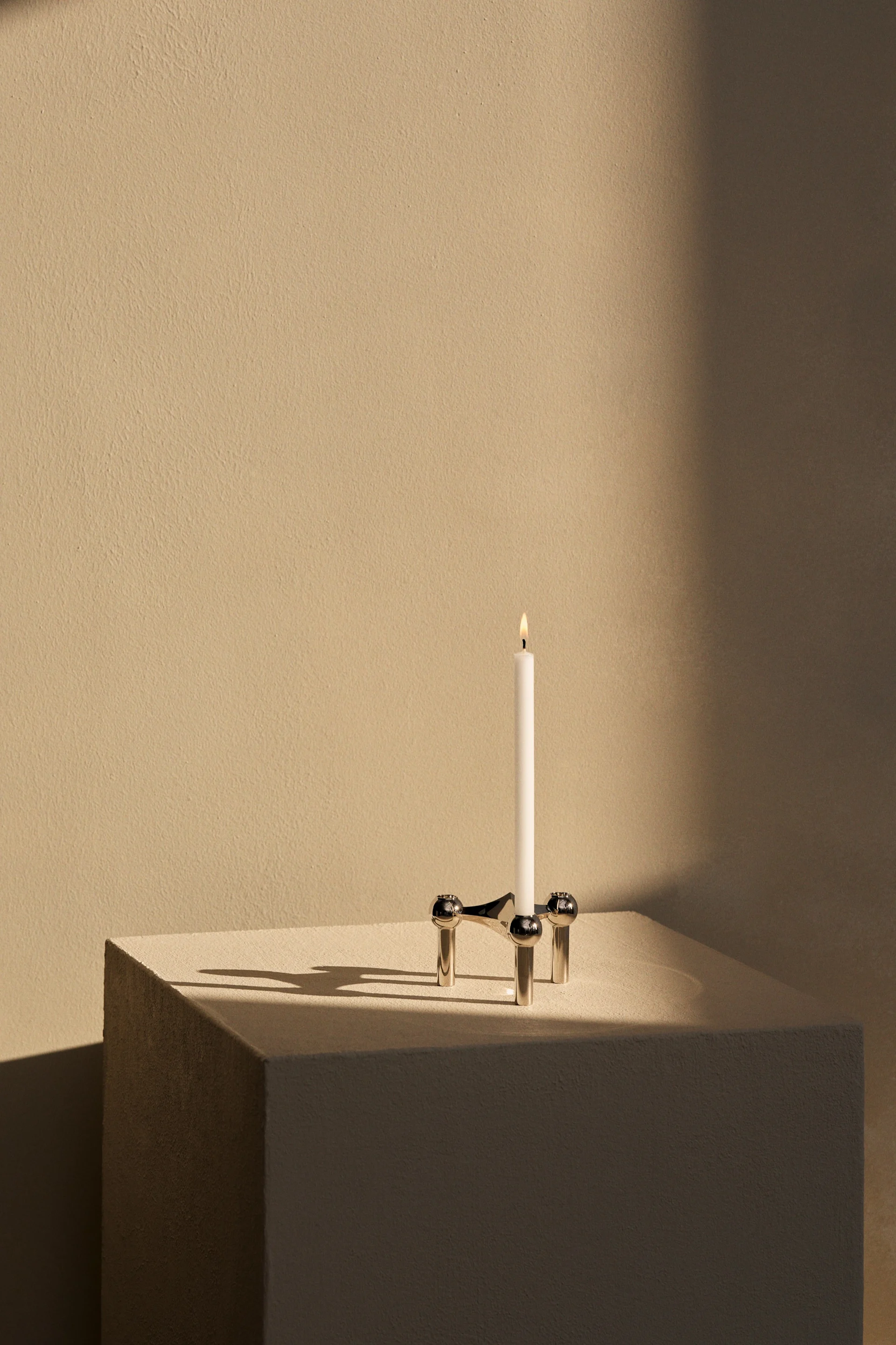 Witte kaars brandt in zilveren, driebenige kandelaar met bolvormige details, schaduwen op lichtbeige ondergrond.
