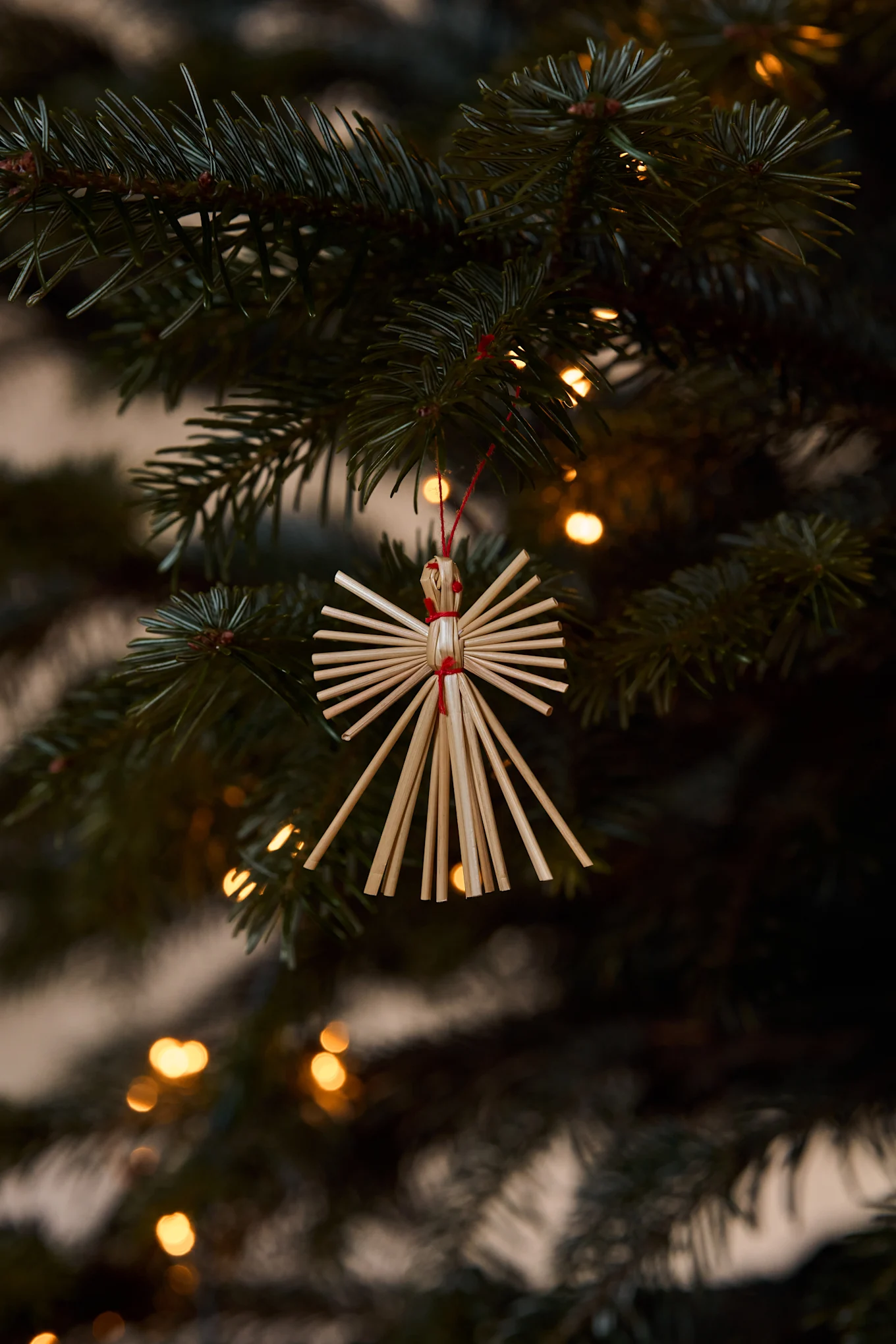 Stro-engelornament hangt aan een kerstboomtak met wazige lichtjes op de achtergrond.