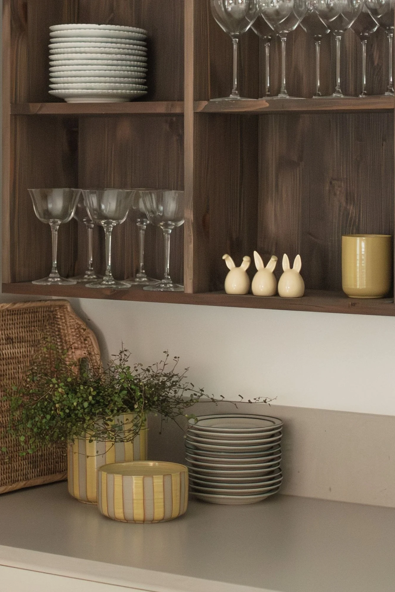 Houten planken met gestapelde witte borden, diverse glazen, drie keramische konijntjes en een gele mok. Op het aanrecht staan een gevlochten mand, planten in gestreepte potten en meer borden.