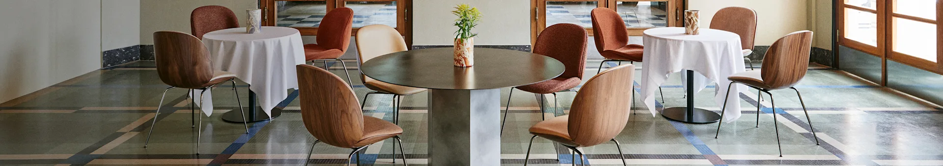 Stijlvol café-interieur met een grote centrale tafel, twee kleinere tafels met witte kleden en diverse stoelen op een gedessineerde vloer.