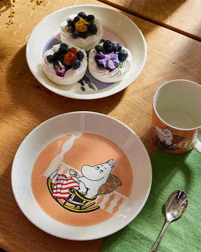 Moomintroll bord, mok en lepel naast een bord met meringues met bosbessen en bloemen op een houten tafel.