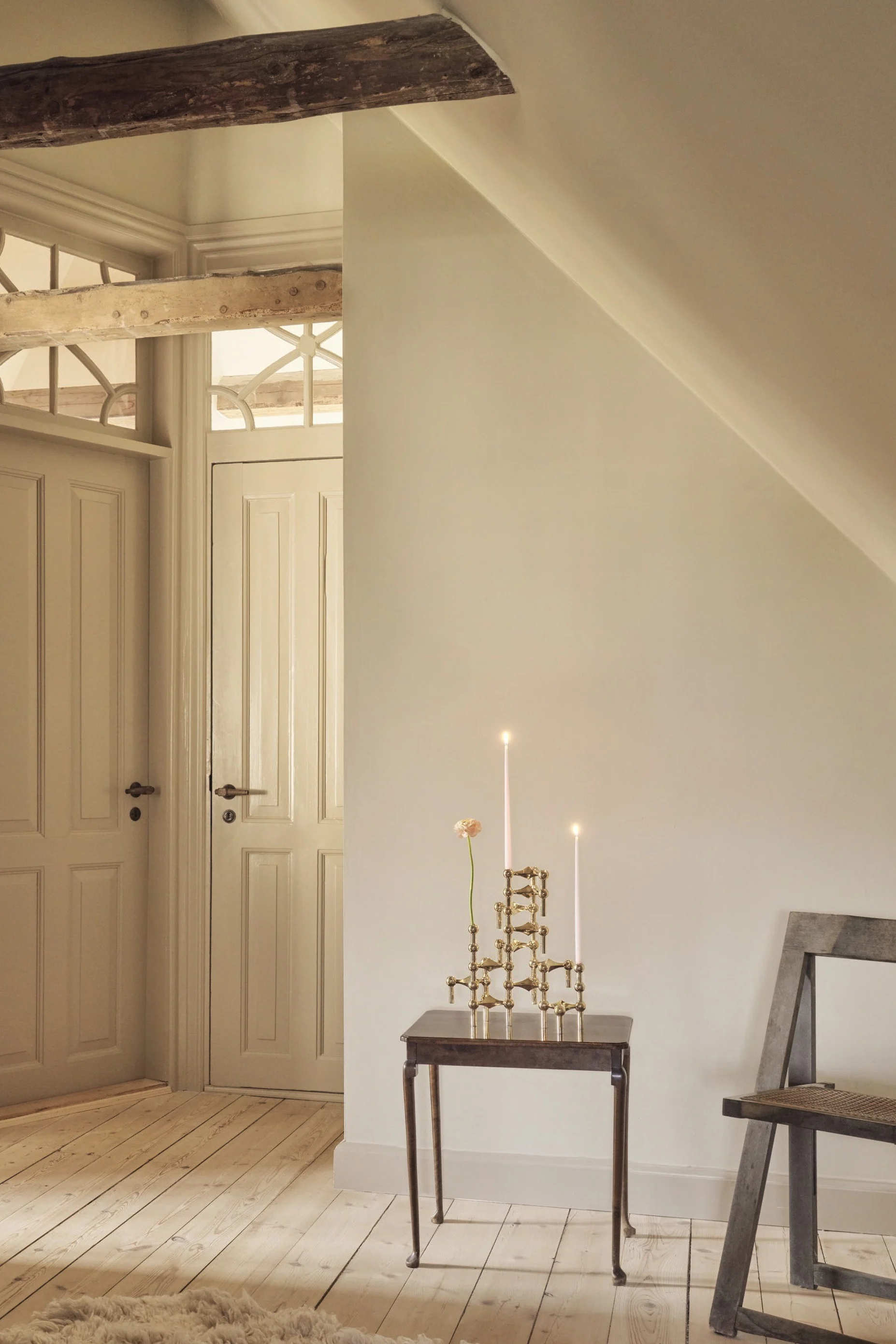 Een zolderkamer met lichte muren en houten vloeren. Twee paneeldeuren met sierlijke bovenlichten, een zichtbare balk, een bijzettafeltje met brandende kaarsen en een bloem, een stoel en een pluizig tapijt.