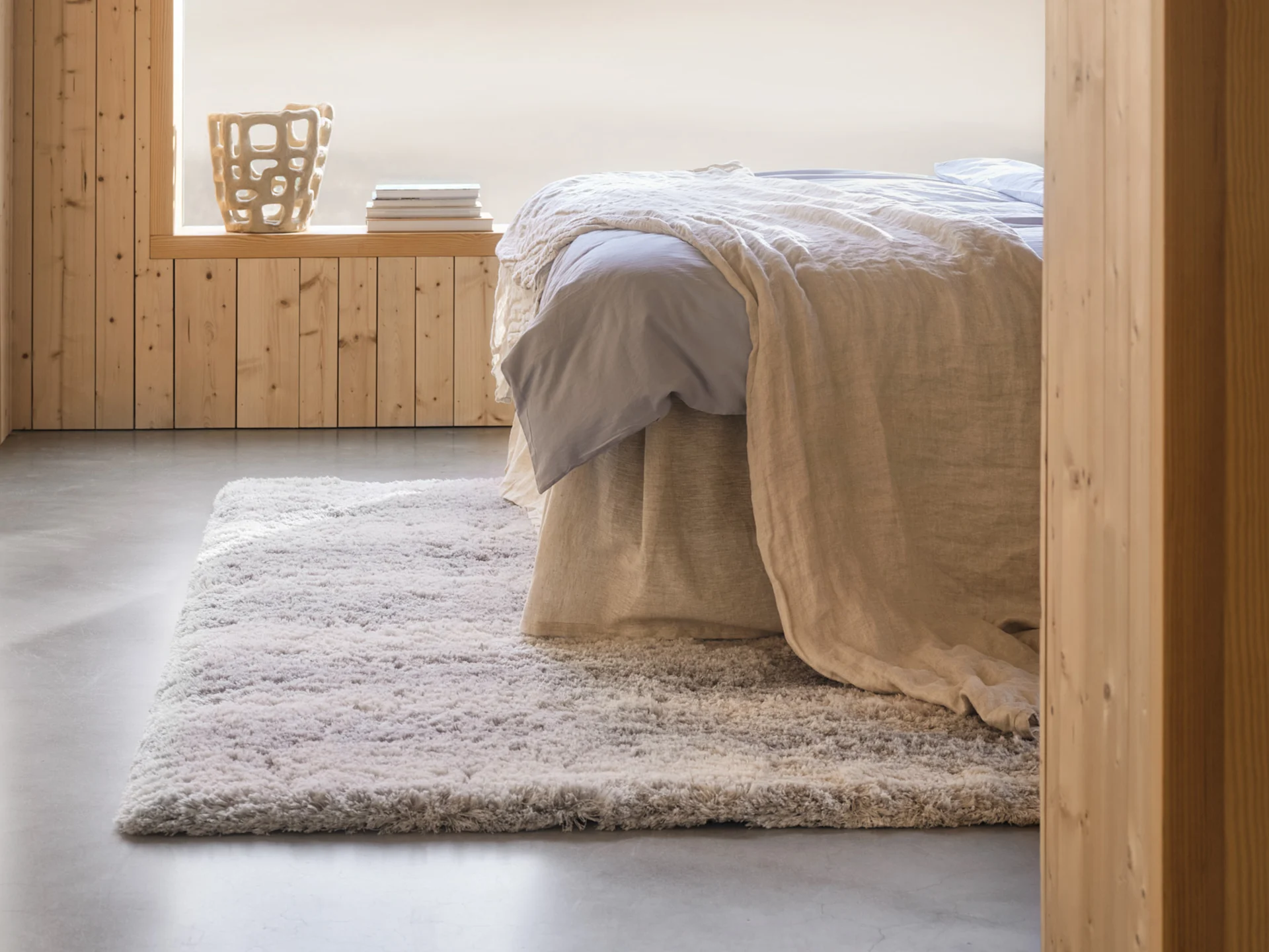 Gezellige slaapkamer met een bed, lichtblauwe lakens, beige deken, een shaggy tapijt, houten lambrisering en een decoratieve vaas.