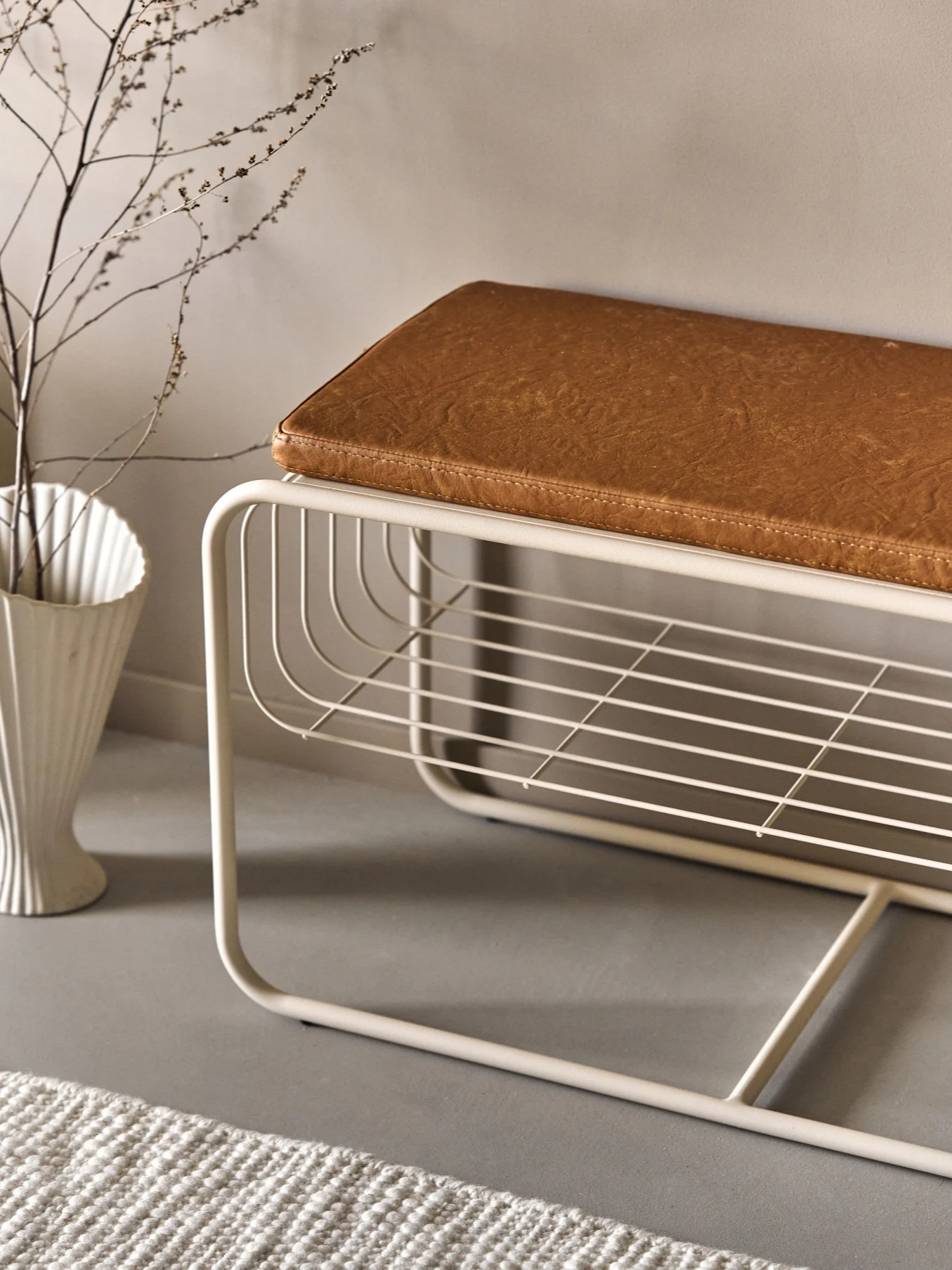 Moderne beige metalen bank met bruin imitatieleer kussen en een open lattenplank. Een witte vaas met takken en een vloerkleed zijn in de buurt.