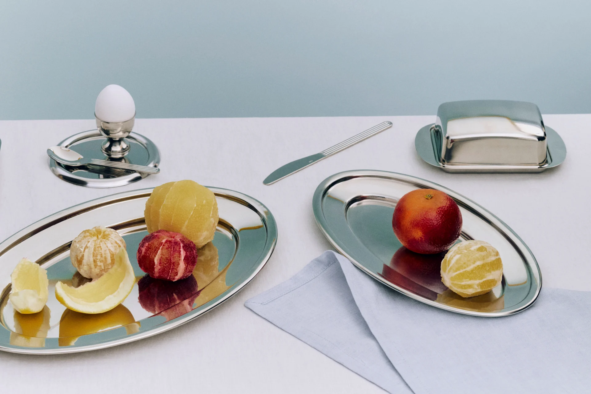 Een ontbijtopstelling met twee zilveren schalen met geschilde citrusvruchten, een ei in een eierdop, een botervloot en een mes.