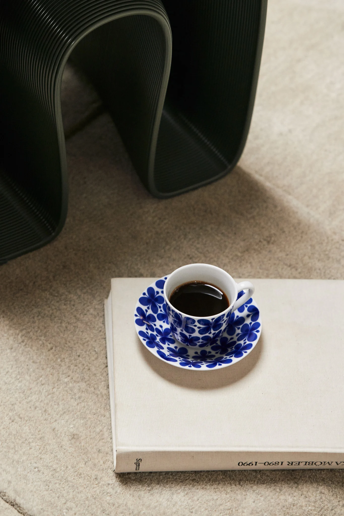 Kopje koffie met blauw bloemen schoteltje op een licht boek, donker geribbeld object op de achtergrond.