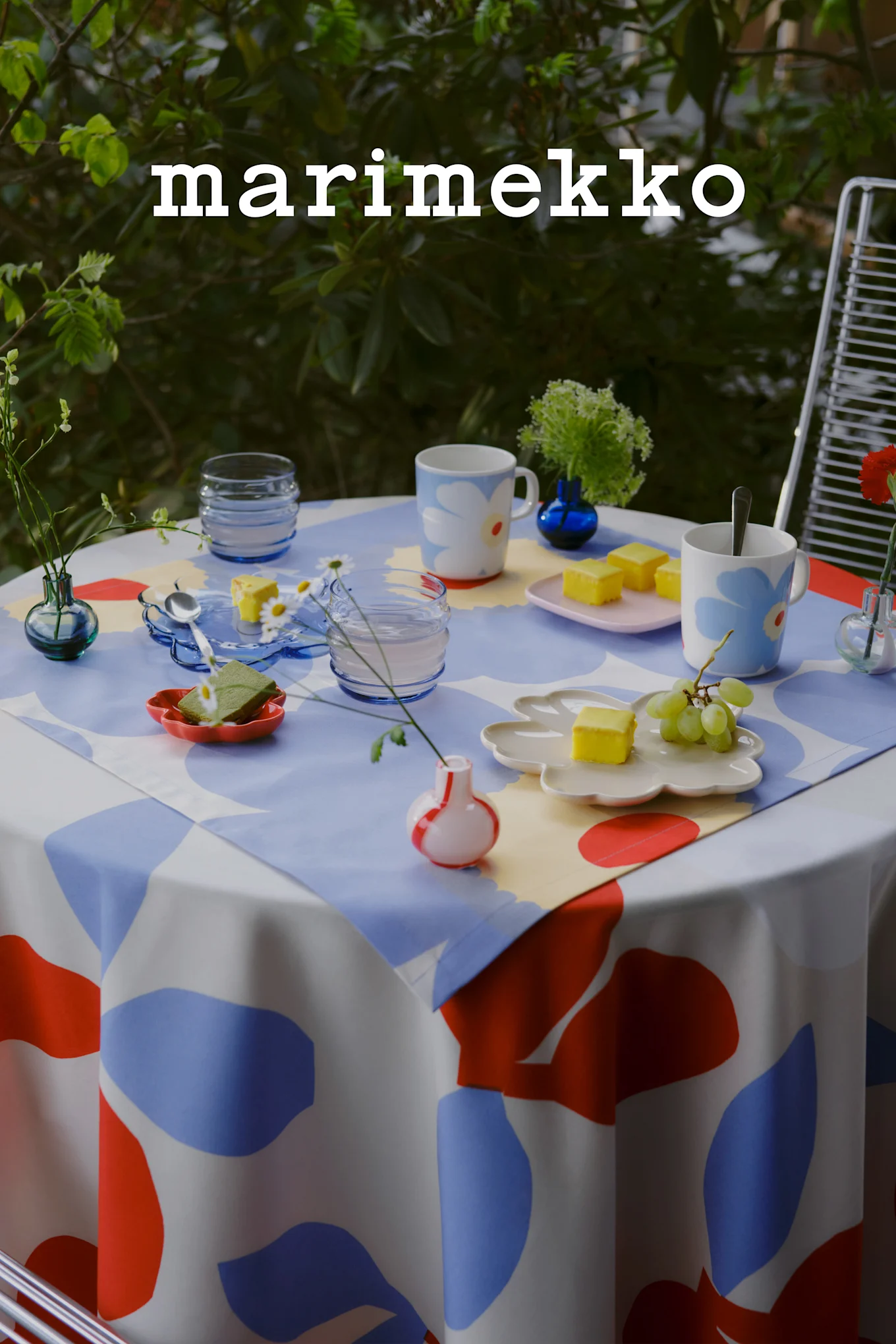 Marimekko-logo. Buitentafel gedekt met blauw, rood en wit abstract patroon tafelkleed, bloemige mokken, gele cakes, druiven en kleine vazen.