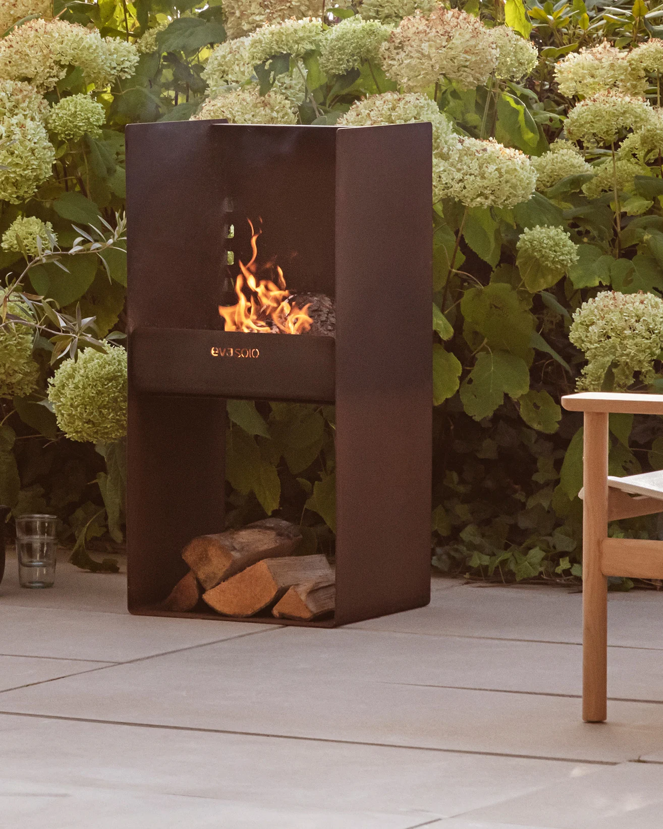 Moderne donkere metalen buitenhaard met brandend vuur, gestapelde houtblokken eronder, omringd door groene planten en een terras.