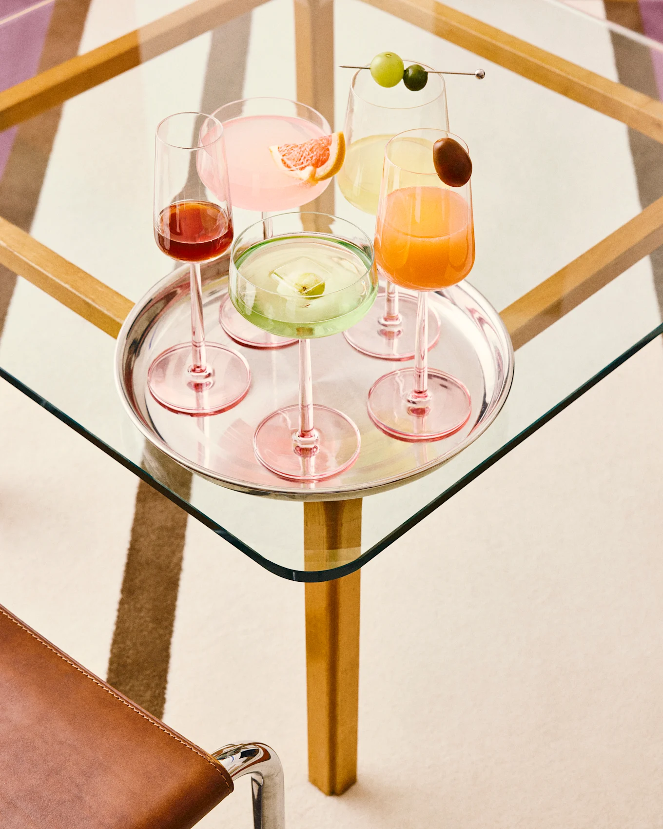 Vijf kleurrijke cocktails op een zilveren dienblad: roze met grapefruit, groen met fruitijs, geel met druiven, oranje met een garnering en donkerrood, allemaal op een glazen tafel.