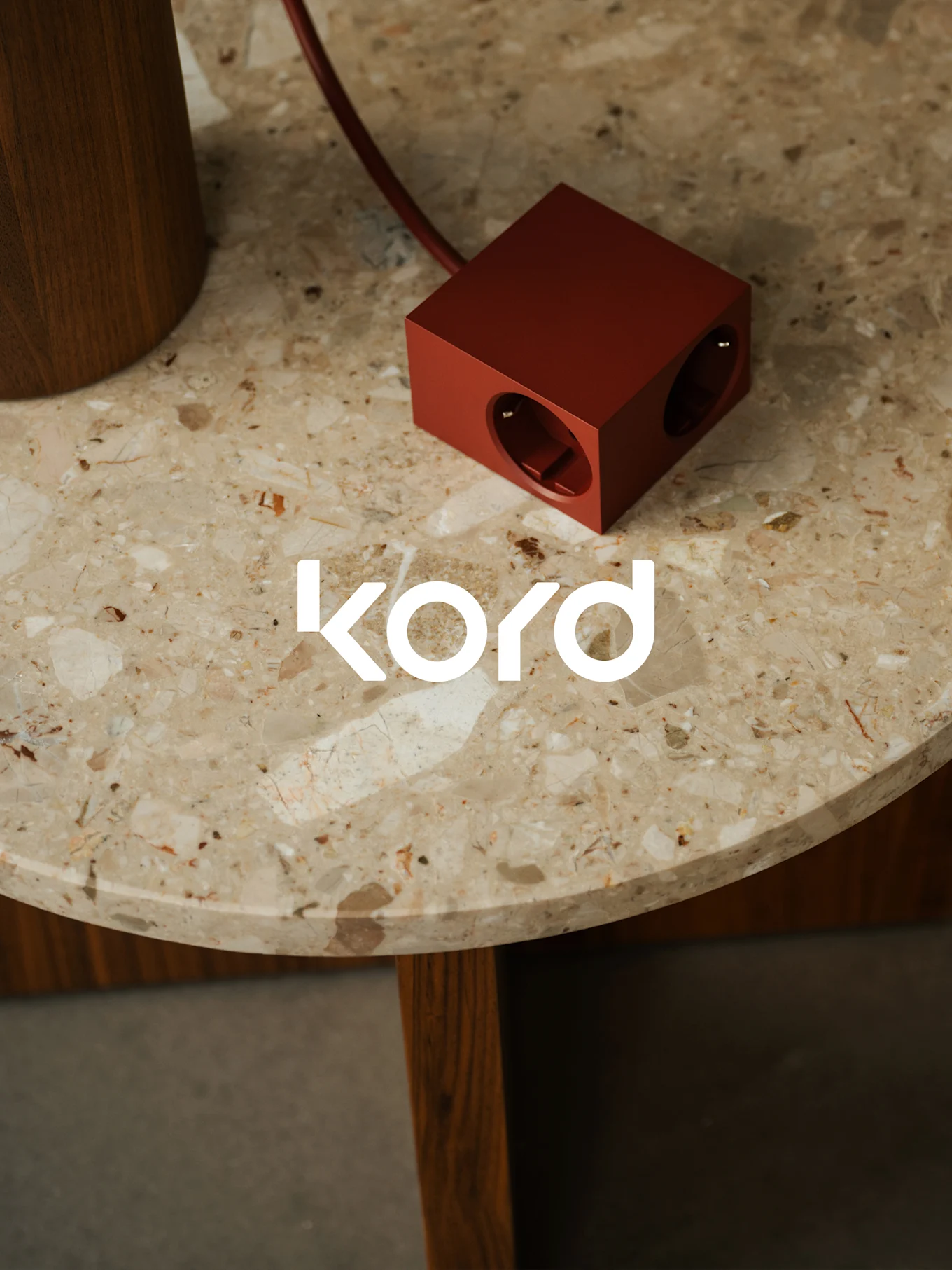 Een donkerrode, kubusvormige stekkerdoos met twee zichtbare stopcontacten rust op een ronde marmeren tafel, met het witte "kord"-logo.