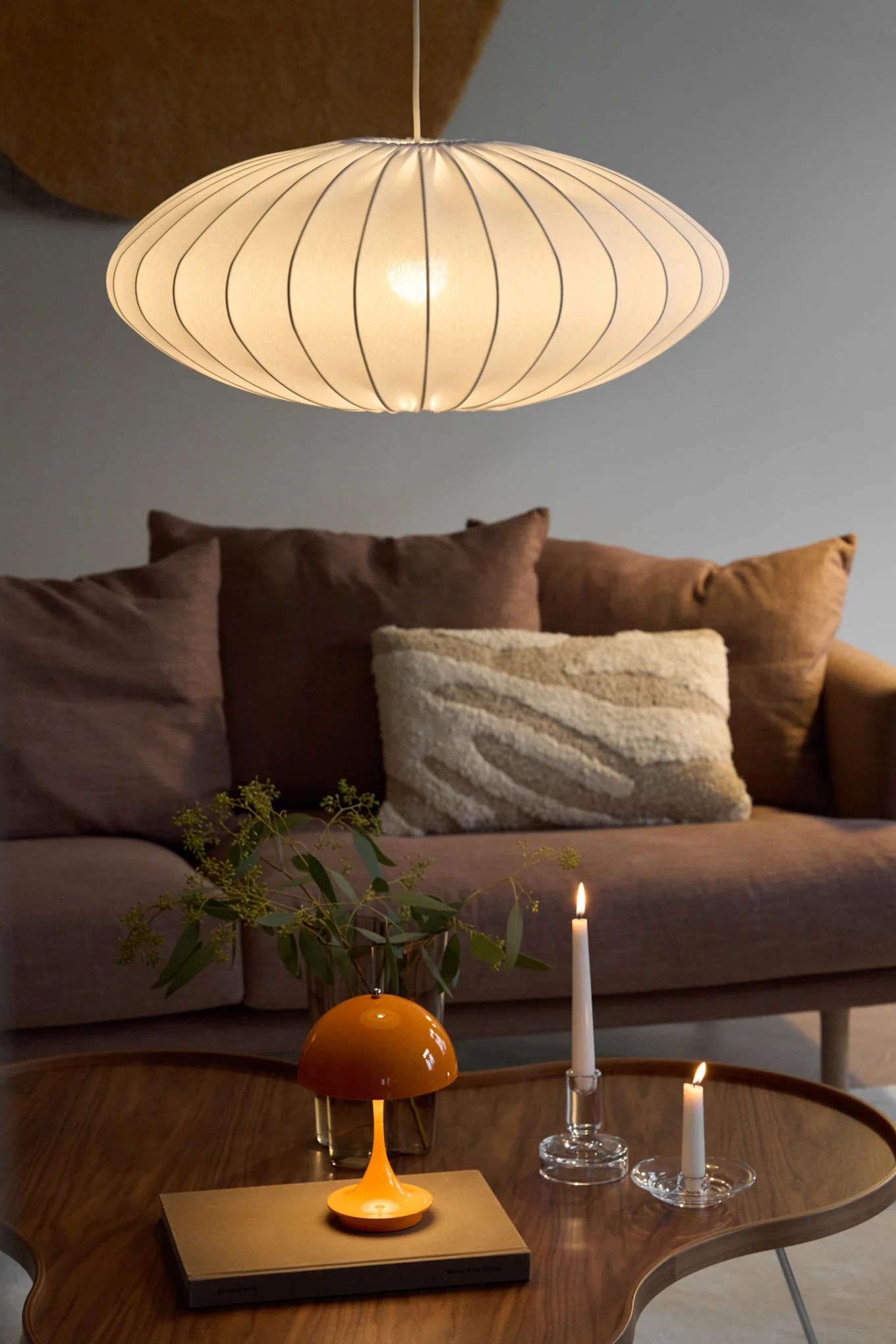 Een ovale, geribbelde hanglamp verlicht een woonkamer met een bruine bank, een houten salontafel, een oranje paddestoellamp en twee brandende kaarsen.