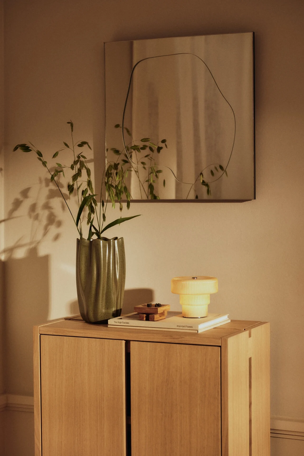 Een houten kast met groene golvende vaas met gras, gele gelaagde lamp en spiegel met onregelmatige omtrek aan de muur.