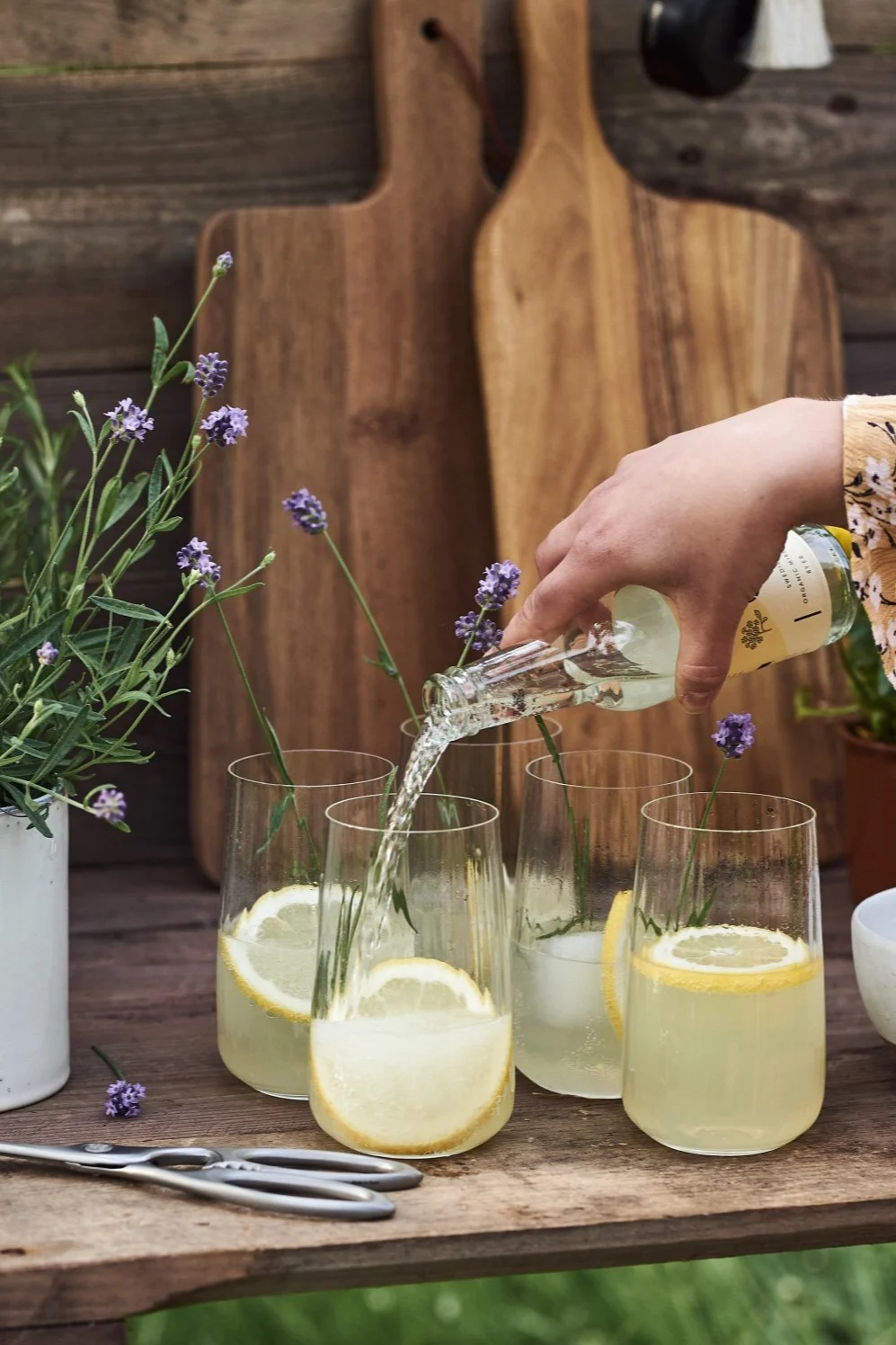 Hand schenkt drank uit kleine fles in glazen met citroenschijfjes en lavendel op een houten tafel.