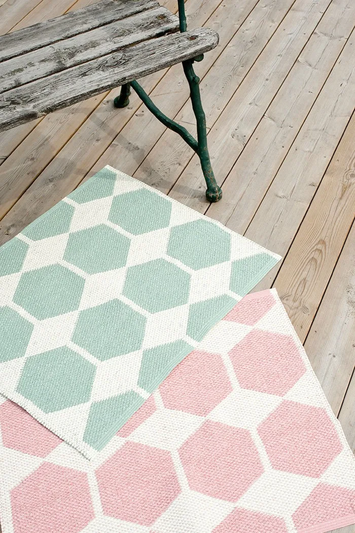 Vloerkleed voor buiten: Hier zie je het groene en roze vloerkleed van Brita Zweden voor een bankje op een houten terras liggen.