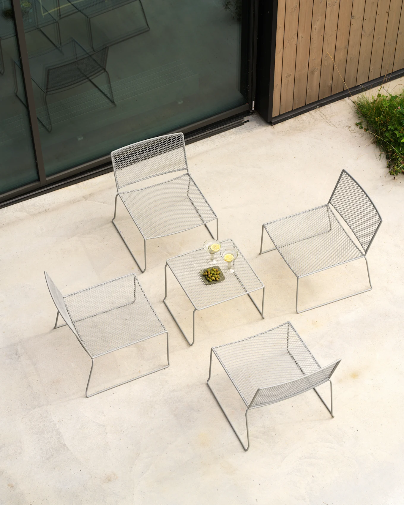 Bovenaanzicht: lichtgrijze buitenloungeset met vier mesh stoelen en tafel met drankjes en olijven op betonnen terras.