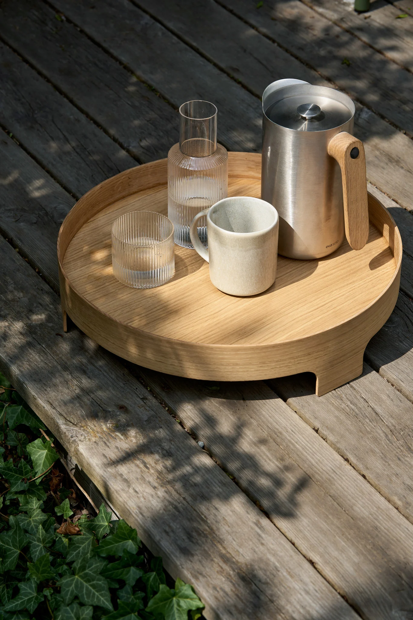 Een rond houten dienblad op een terras met een roestvrijstalen kan met houten handvat, een geribbelde glazen karaf, een geribbeld glas en een keramische mok.