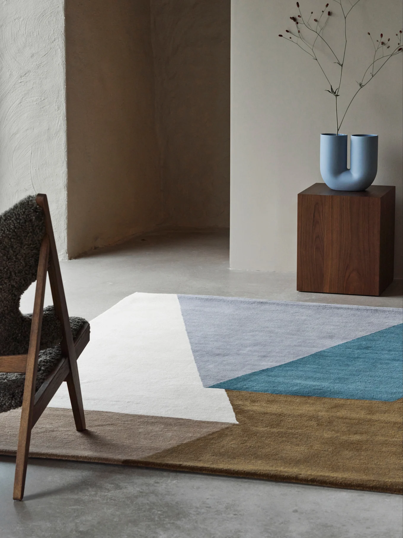 Moderne kamer met een geometrisch vloerkleed in wit, grijs, groenblauw en bruin, een U-vormige blauwe vaas op een houten sokkel en een pluizige stoel.