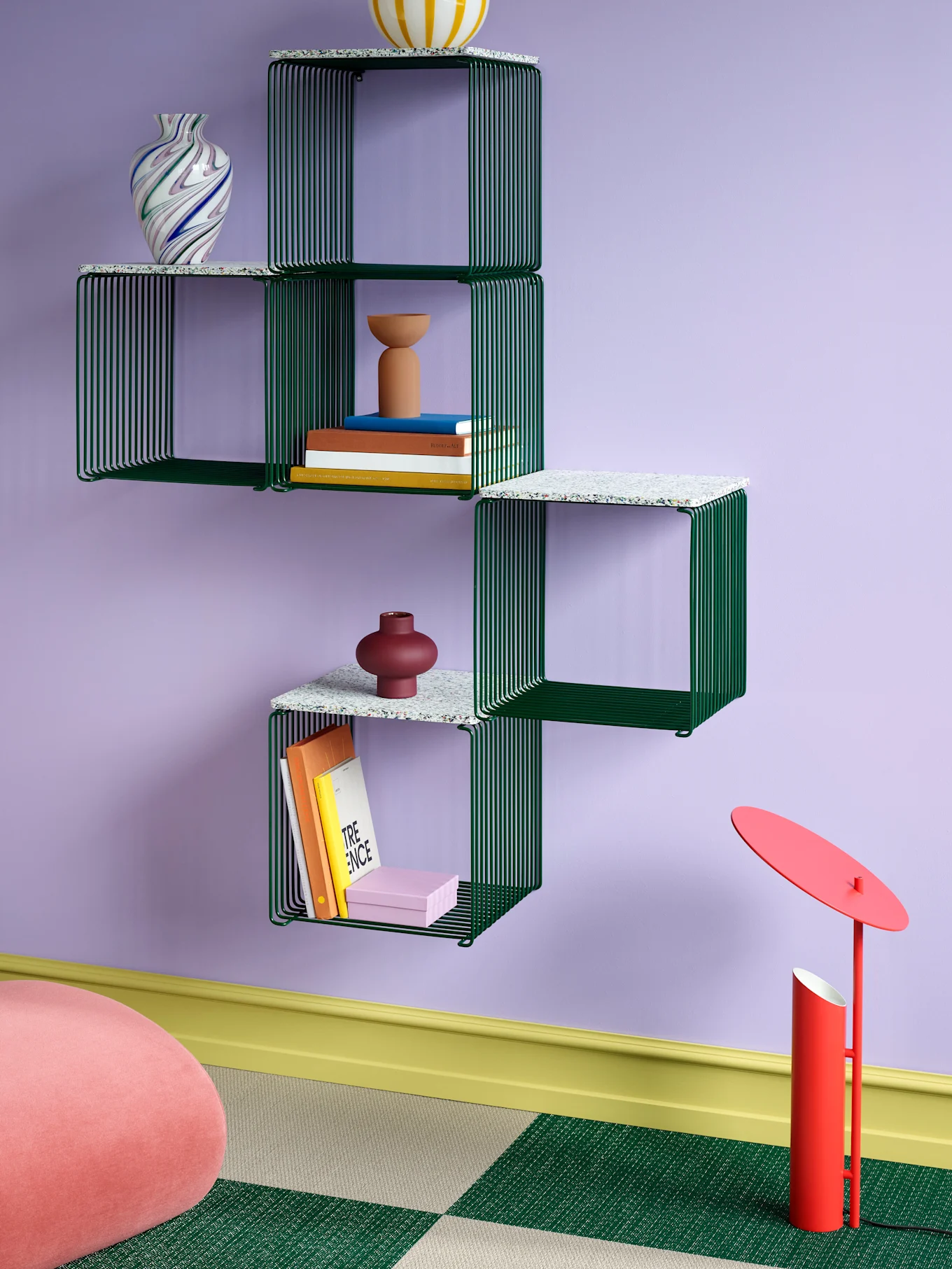 Groene modulaire wandplank op een paarse muur, met vazen, boeken en decoratieve objecten. Een rode vloerlamp en roze poef staan eronder.