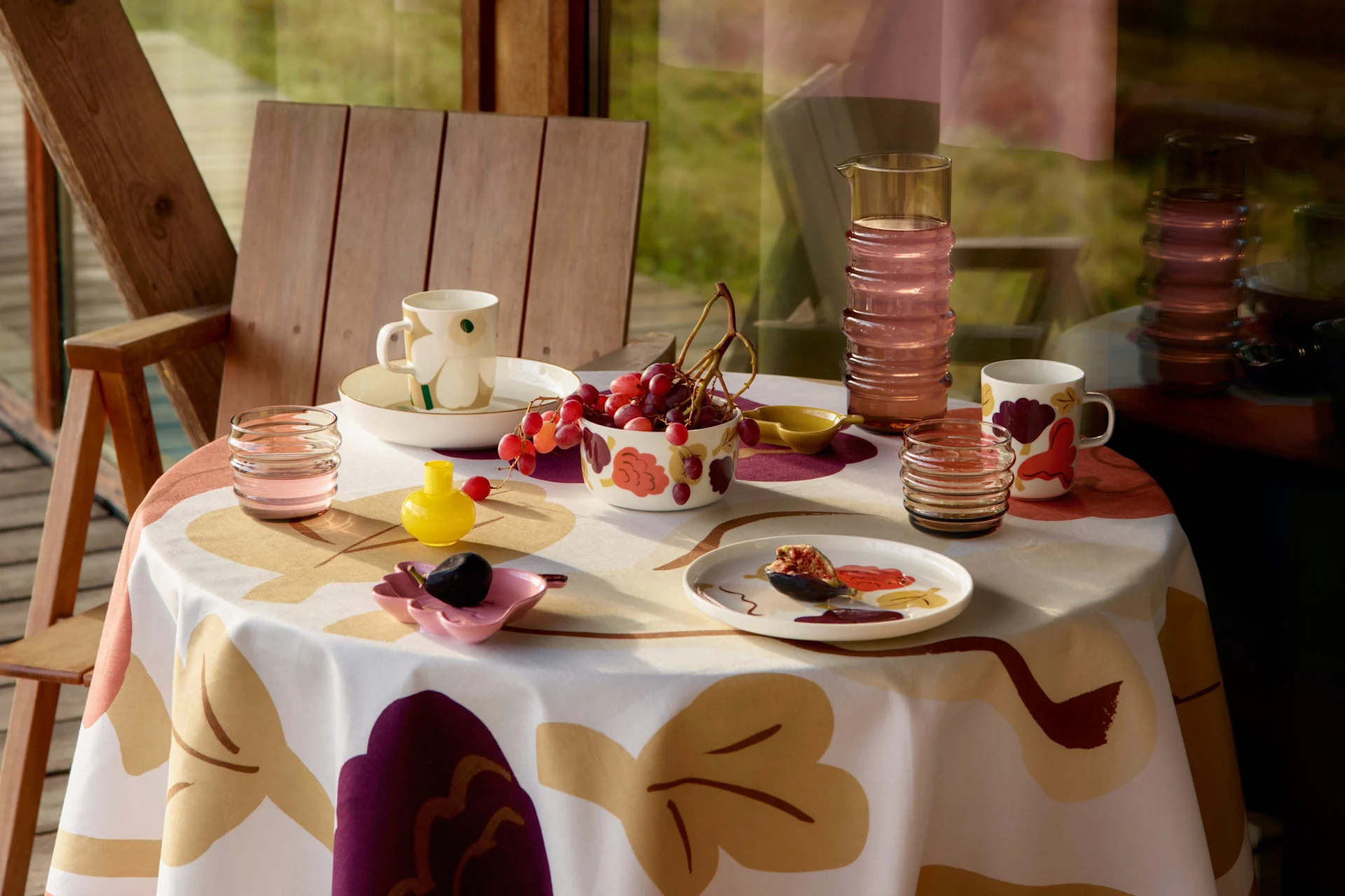 Een tafel gedekt met een gemêleerd tafelkleed, mokken, schalen met druiven, een vijg op een bord, een karaf en glazen, naast een houten stoel.