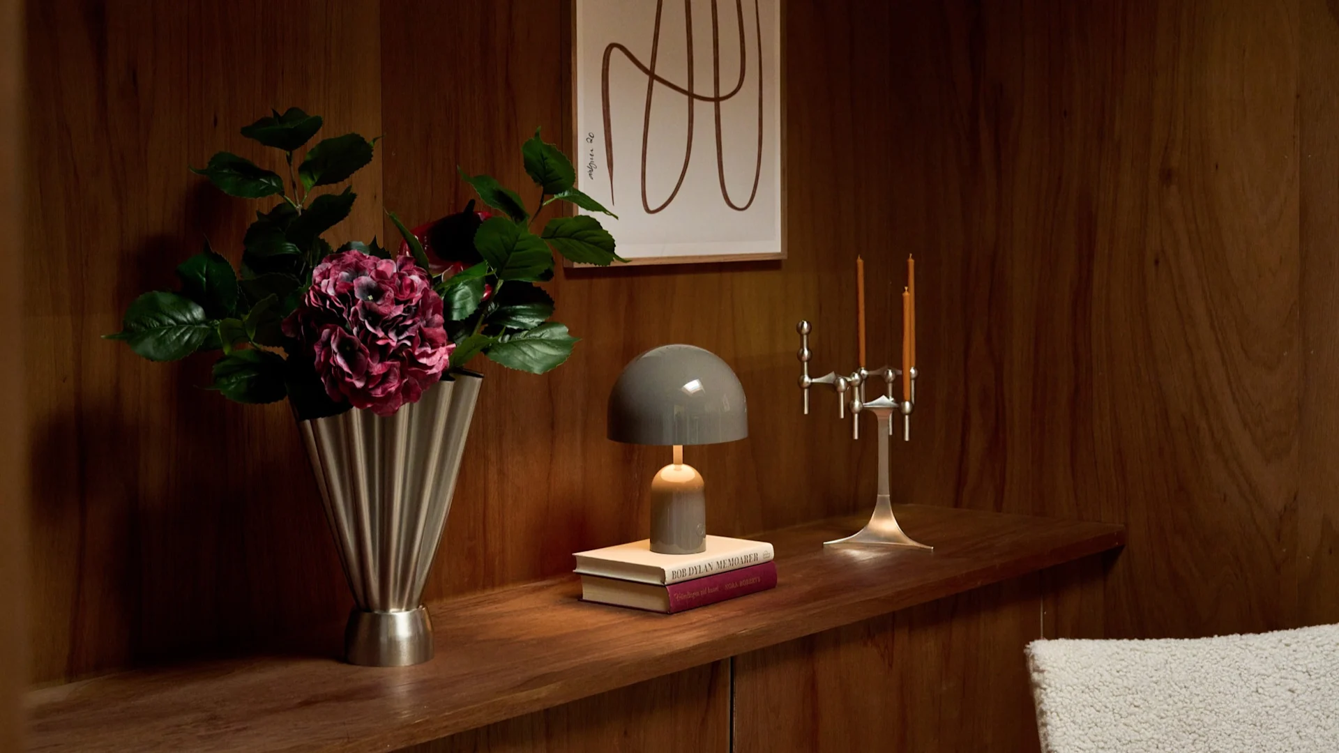 Vaas met donkerroze bloemen, grijze paddenstoellamp op boeken, zilveren kandelaar op een houten plank met kunst.