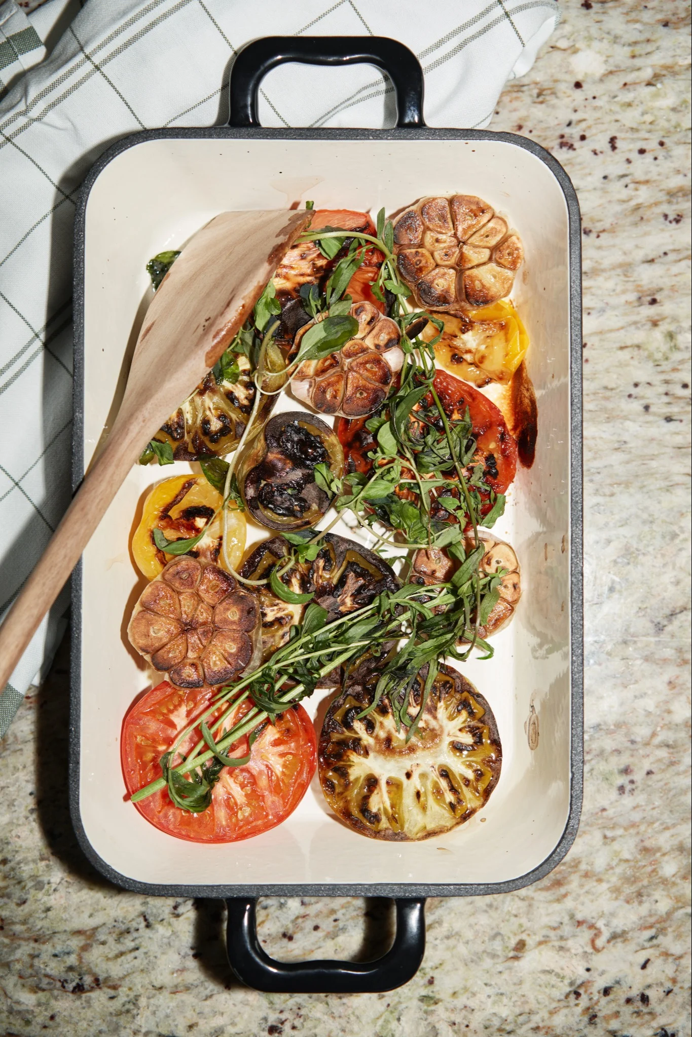 Een witte ovenschaal gevuld met gegrilde tomaten, aubergine, geroosterde knoflook en verse groene kruiden, met een houten lepel.