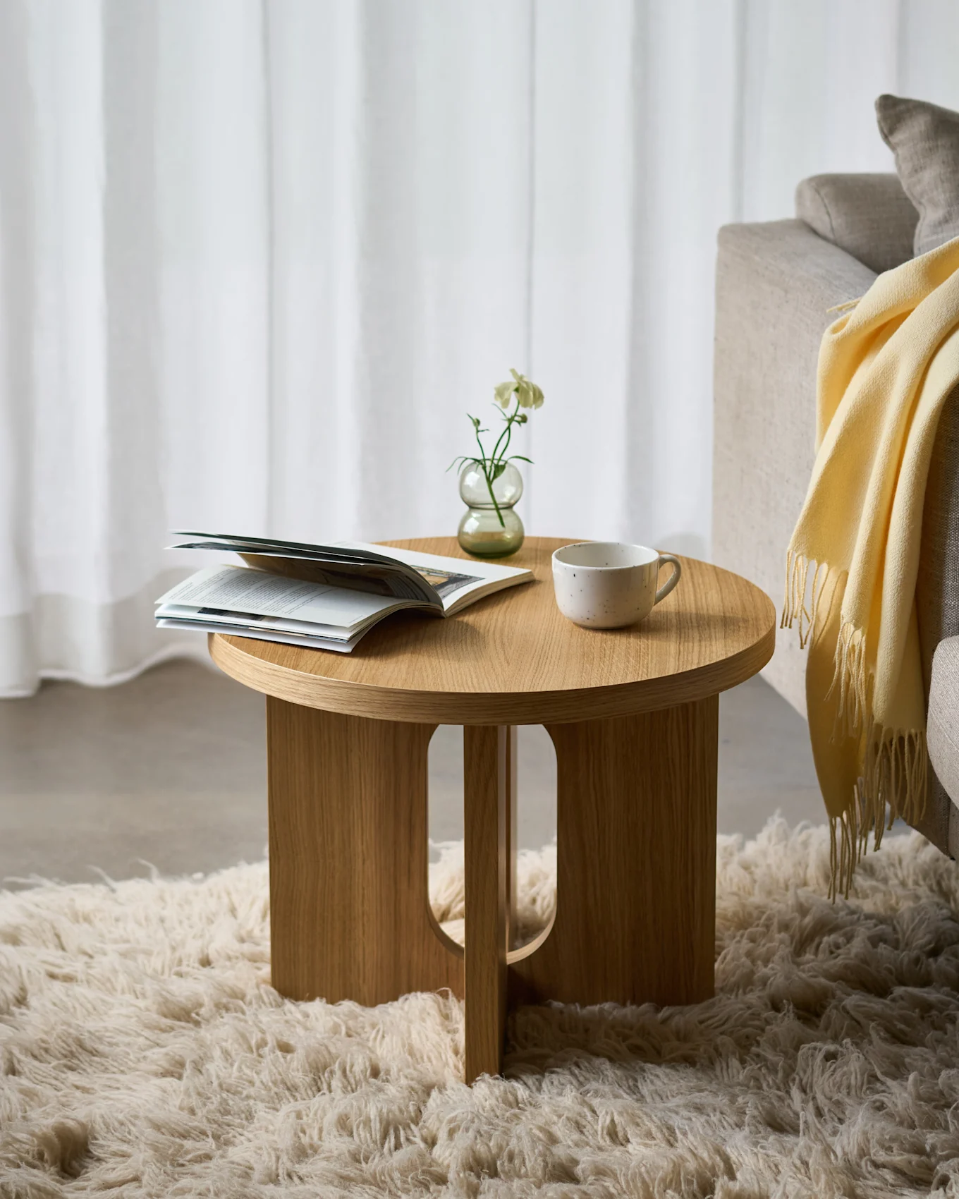 Ronde houten bijzettafel op een pluizig tapijt, met een open tijdschrift, een witte bloem in een vaas en een keramische kop, naast een bank.