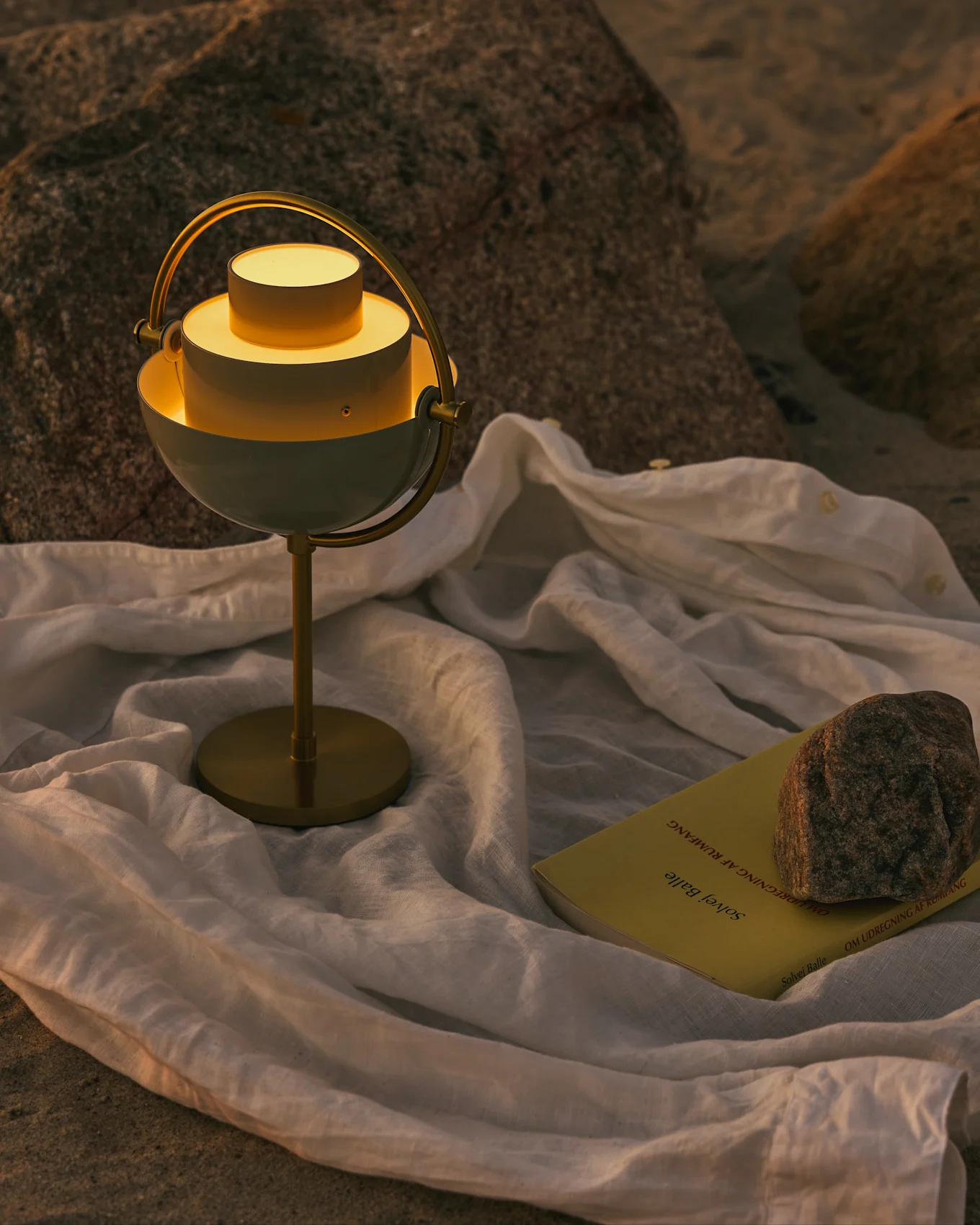 Een moderne wit-messing tafellamp met warm licht, een geel boek en een steen op een witte doek op een rotsachtige ondergrond.