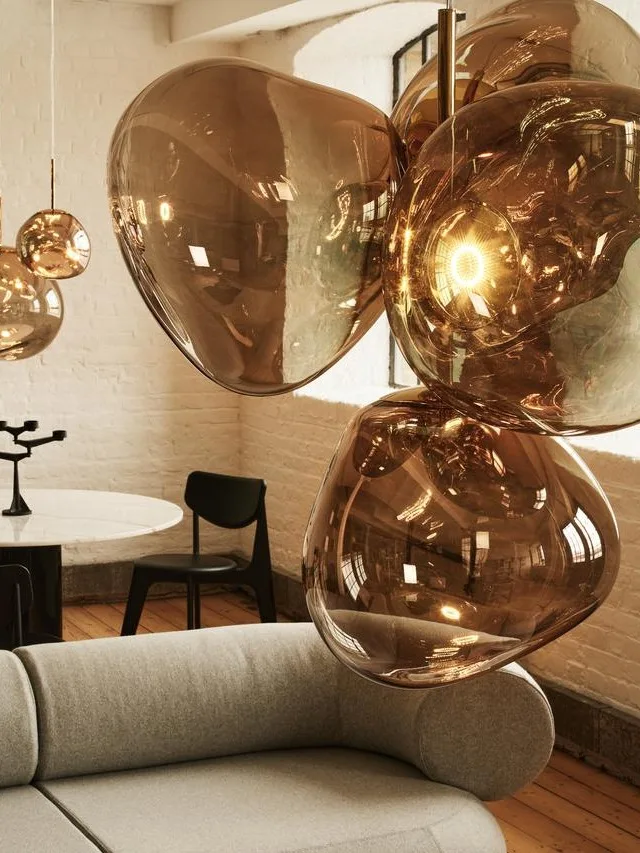 Een moderne woonkamer met een gebogen grijze bank, een bijpassende poef met tijdschriften en amberkleurige glazen hanglampen.