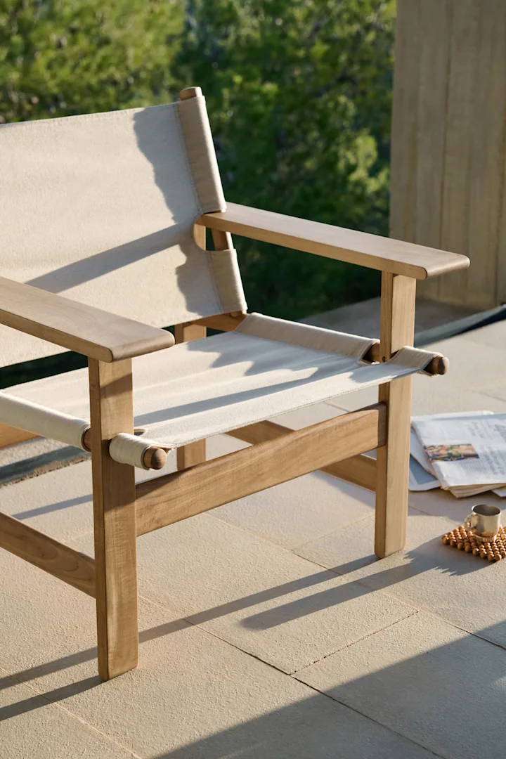 Lichte houten buitenstoel met crèmekleurig canvas op een terras, met een krant en kop ernaast.