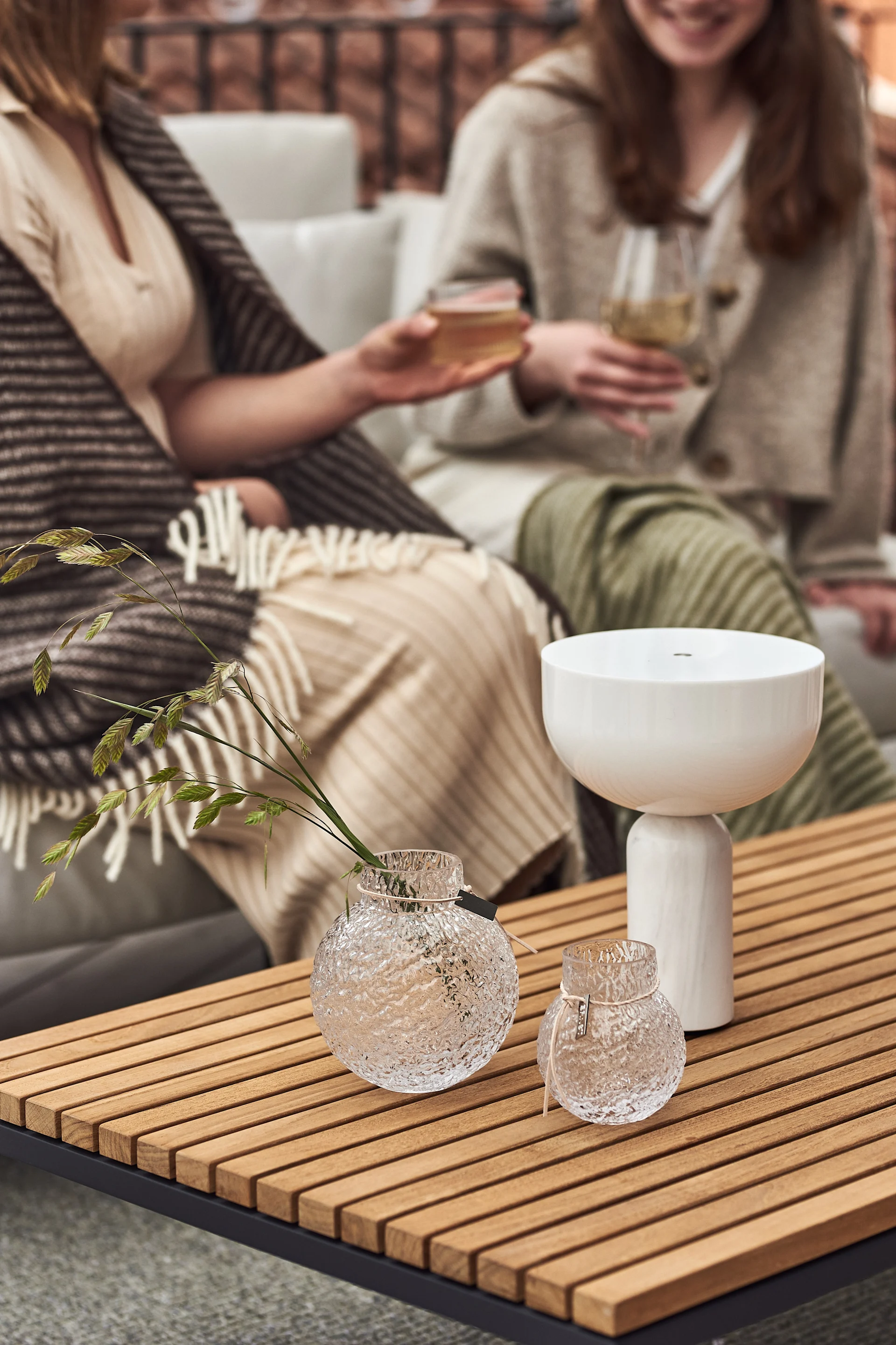 Twee vrouwen zitten op een bank op het balkon en klinken glazen. Op de voorgrond staan de snoerloze Kizu tafellamp van New Works en vazen van Ernst in verschillende maten op een houten tafel.
