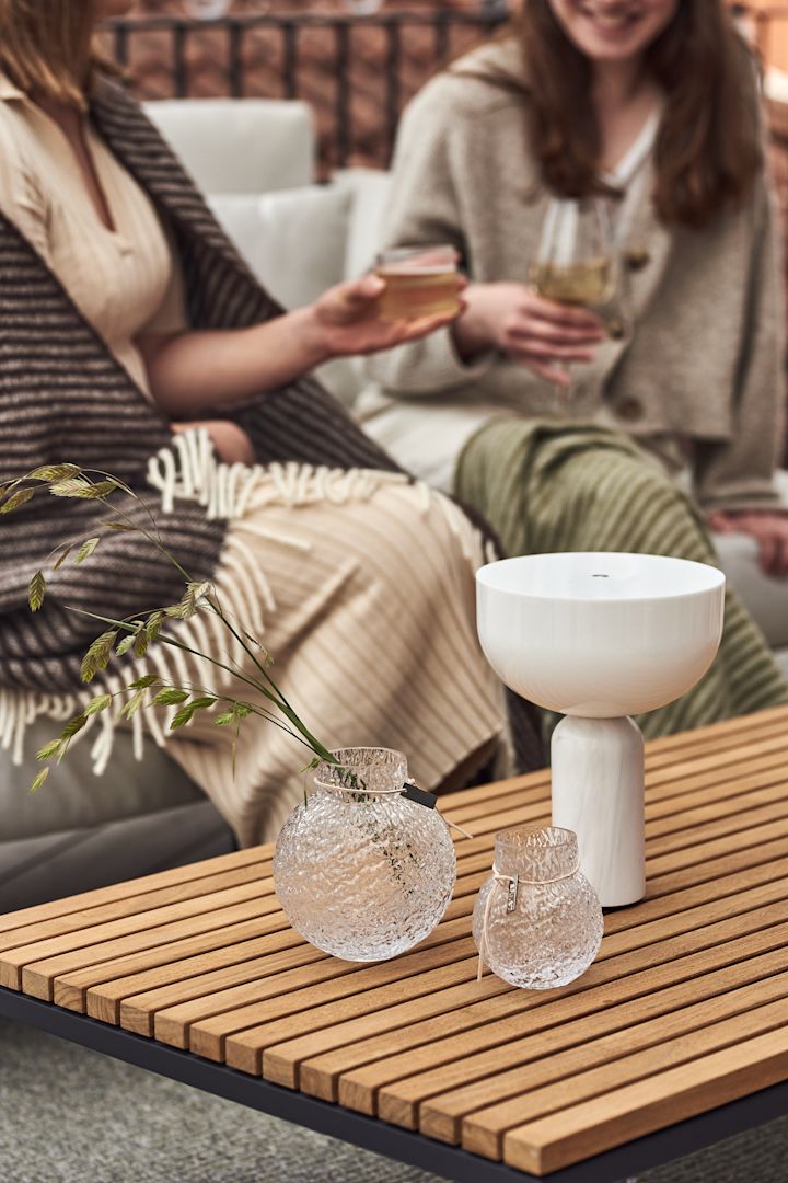 Twee vrouwen zitten op een bank op het balkon en klinken glazen. Op de voorgrond staan de snoerloze Kizu tafellamp van New Works en vazen van Ernst in verschillende maten op een houten tafel.