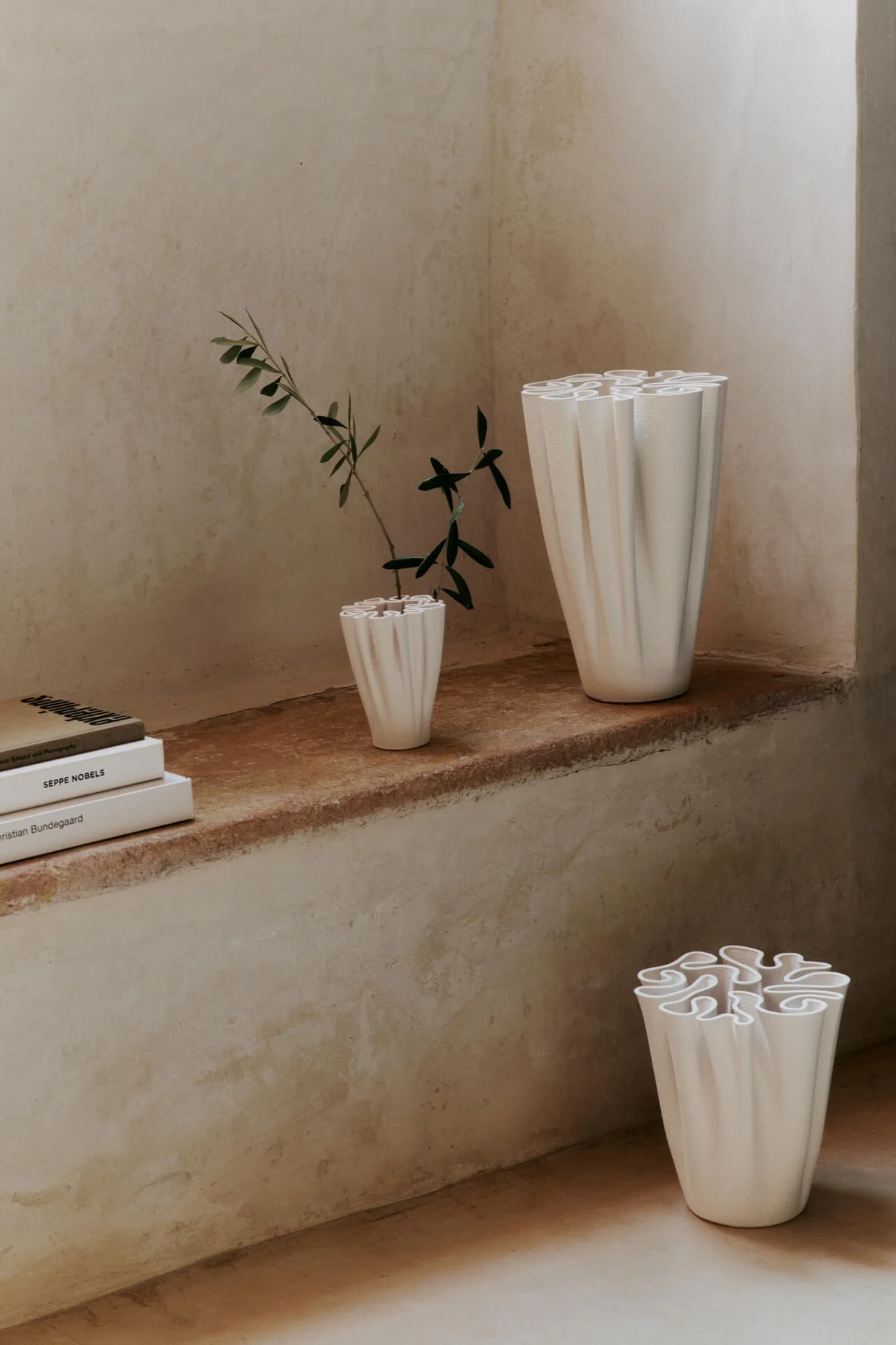 Drie witte, geplooide vazen tentoongesteld in een rustieke kamer, waarvan één met een groene plant.