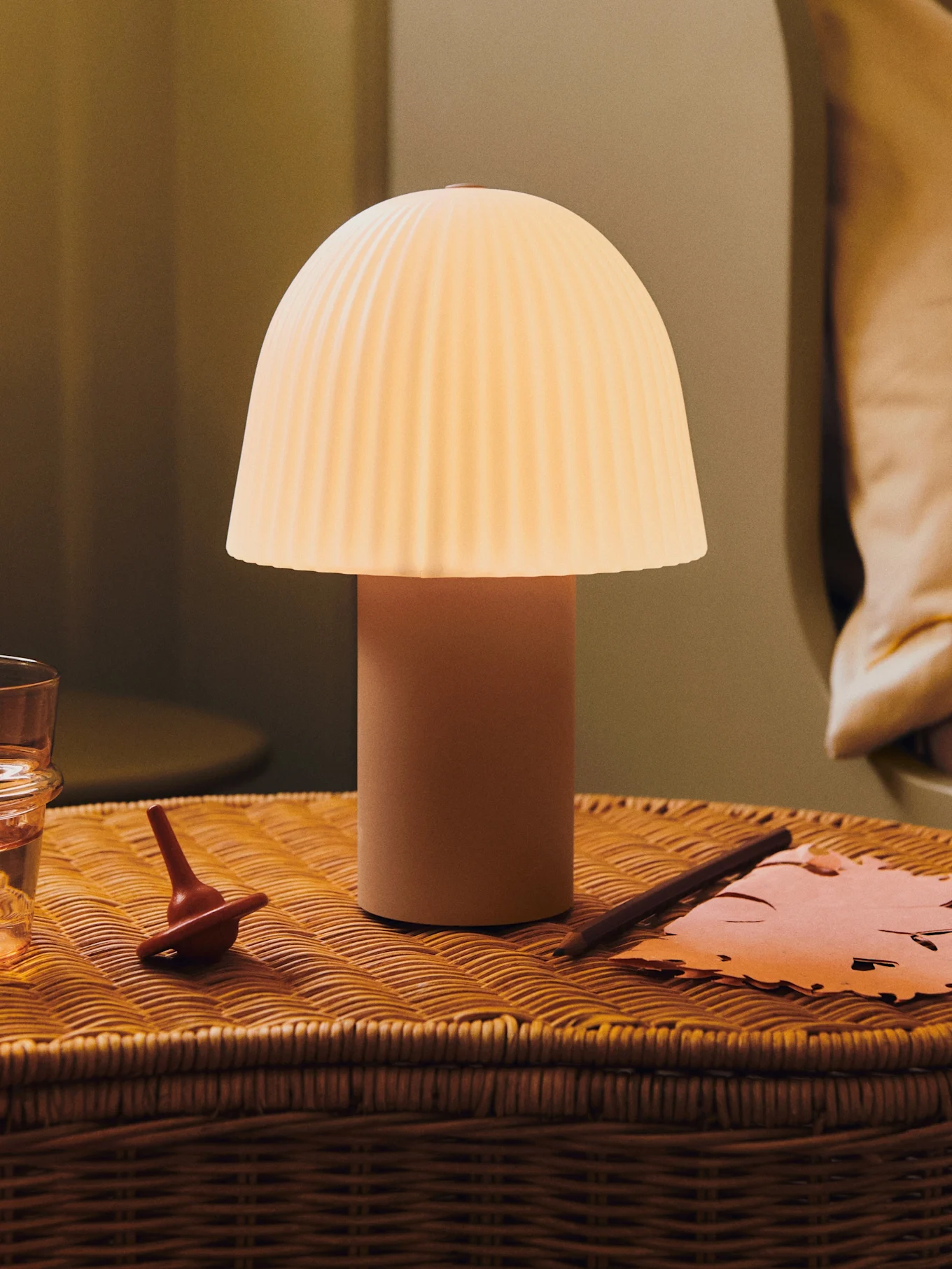 Een gloeiende paddestoellamp met geplooide kap op een rieten nachtkastje met een glas water, een tol en papier.
