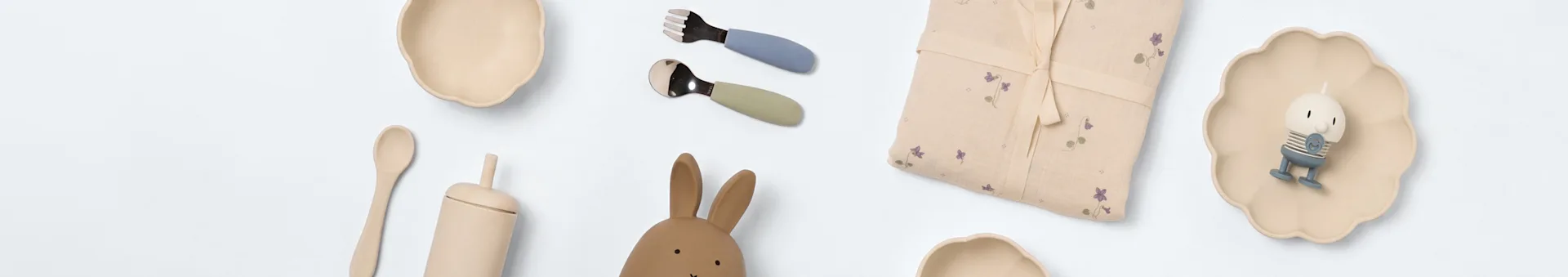 Flat lay van baby eet- en speelgoedartikelen: beige kommen, blauwe vork, groene lepel, bloemendeken, karakterkaars op een bord, houten lepel, konijnenspeelgoed.