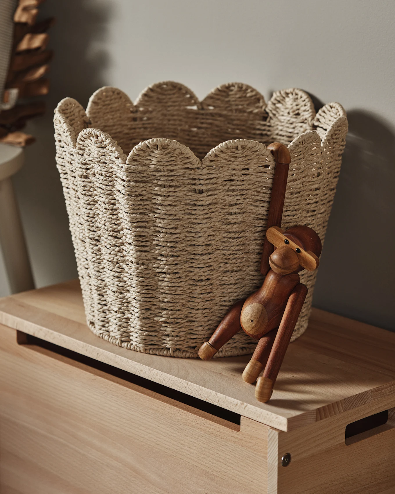 Een lichte gevlochten mand met een geschulpte rand staat op een houten oppervlak, met een houten apenbeeldje dat eraan hangt.