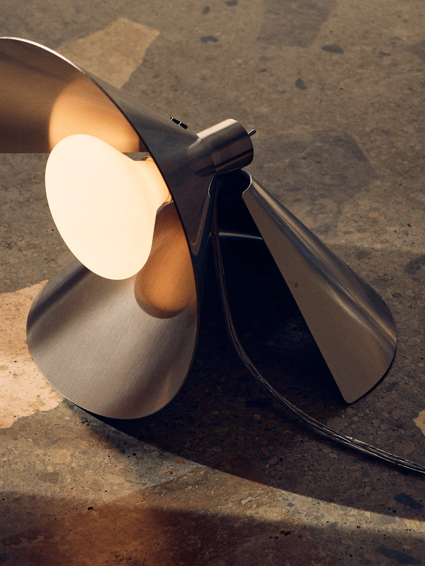 Moderne geborstelde metalen tafellamp met sculpturaal, gevouwen ontwerp en warme, zichtbare gloeilamp op een textuurondergrond.