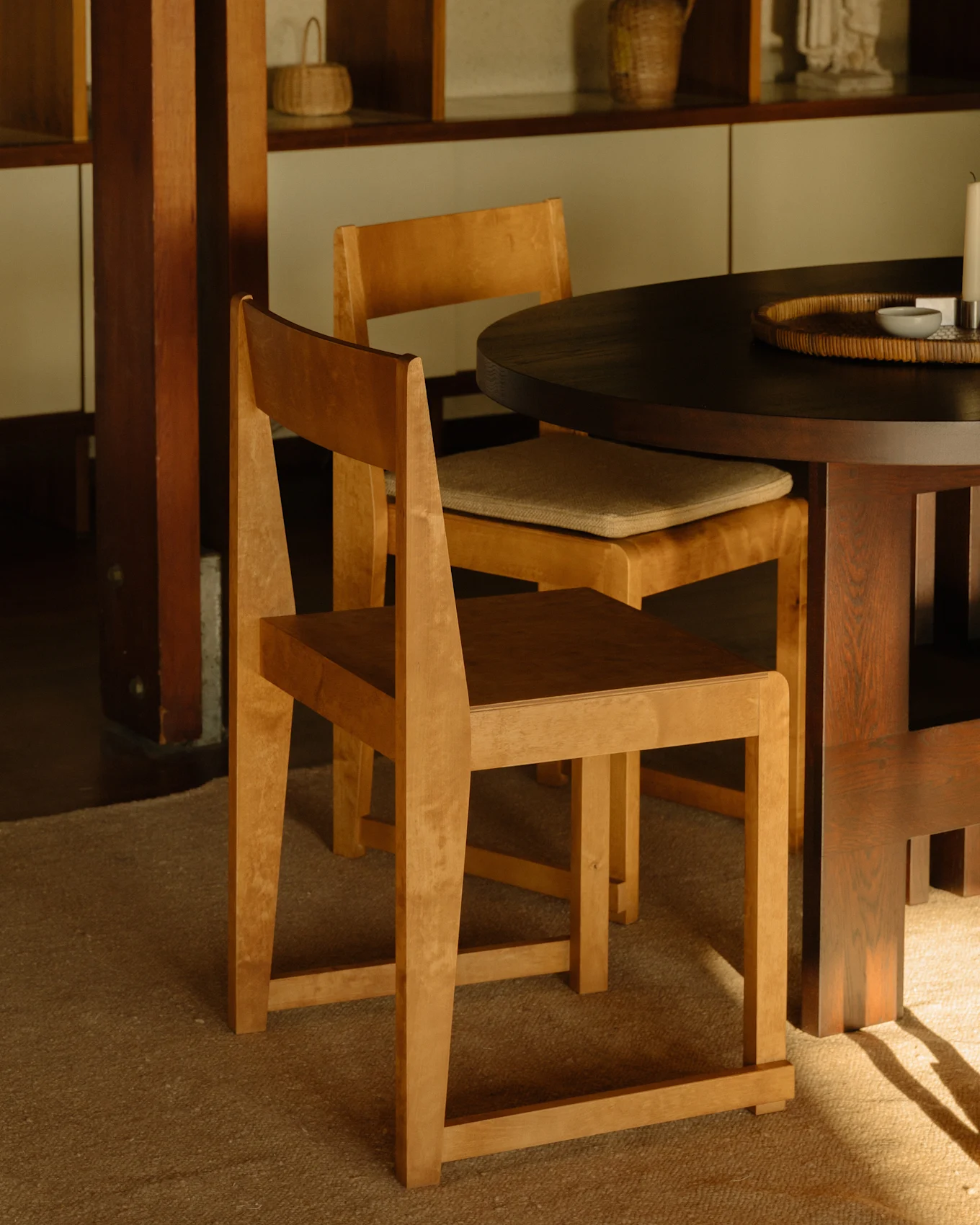 Twee lichte houten stoelen aan een donkere ronde eettafel, met houten planken op de achtergrond.