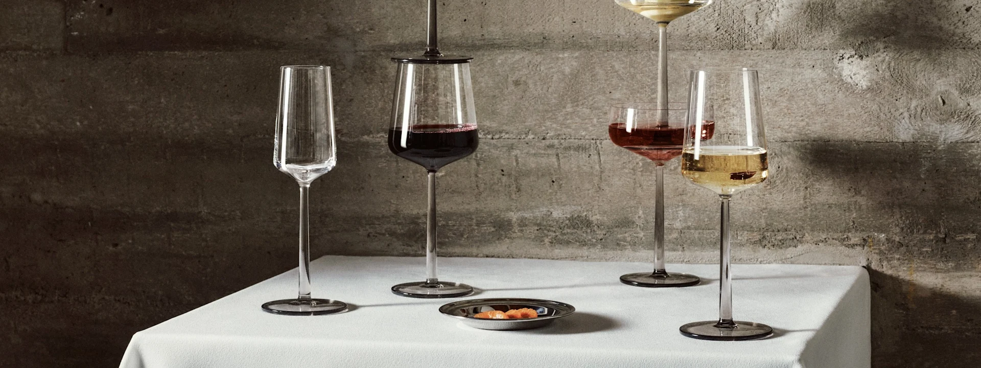 Vijf elegante langstelige glazen met wijn en champagne, één met een peer, op een wit tafelkleed voor een betonnen muur.