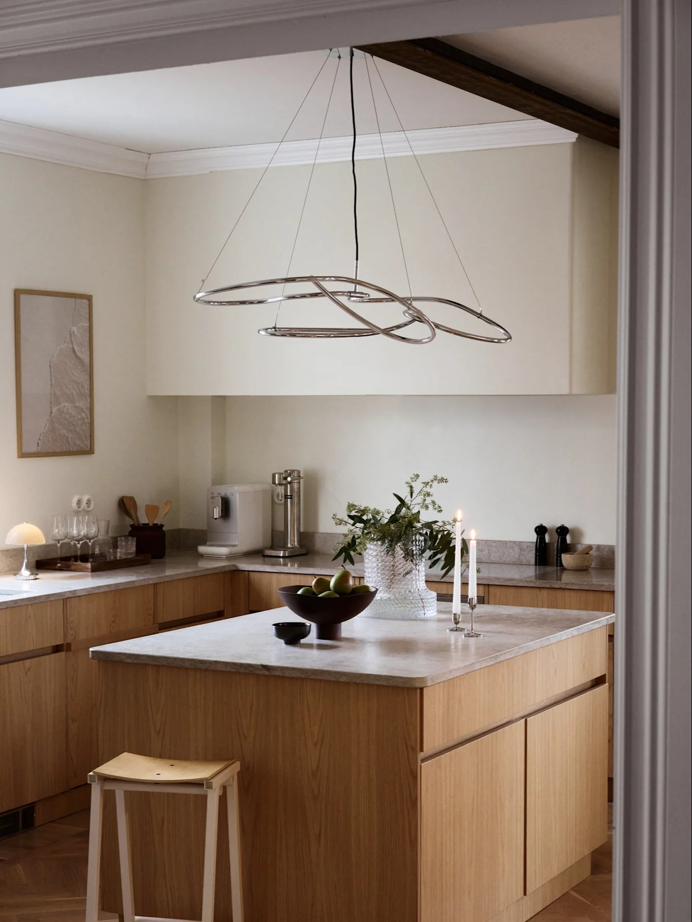Een moderne keuken met lichte houten kasten, grijze aanrechtbladen en een chromen abstracte kroonluchter boven een eiland met fruit en kaarsen.
