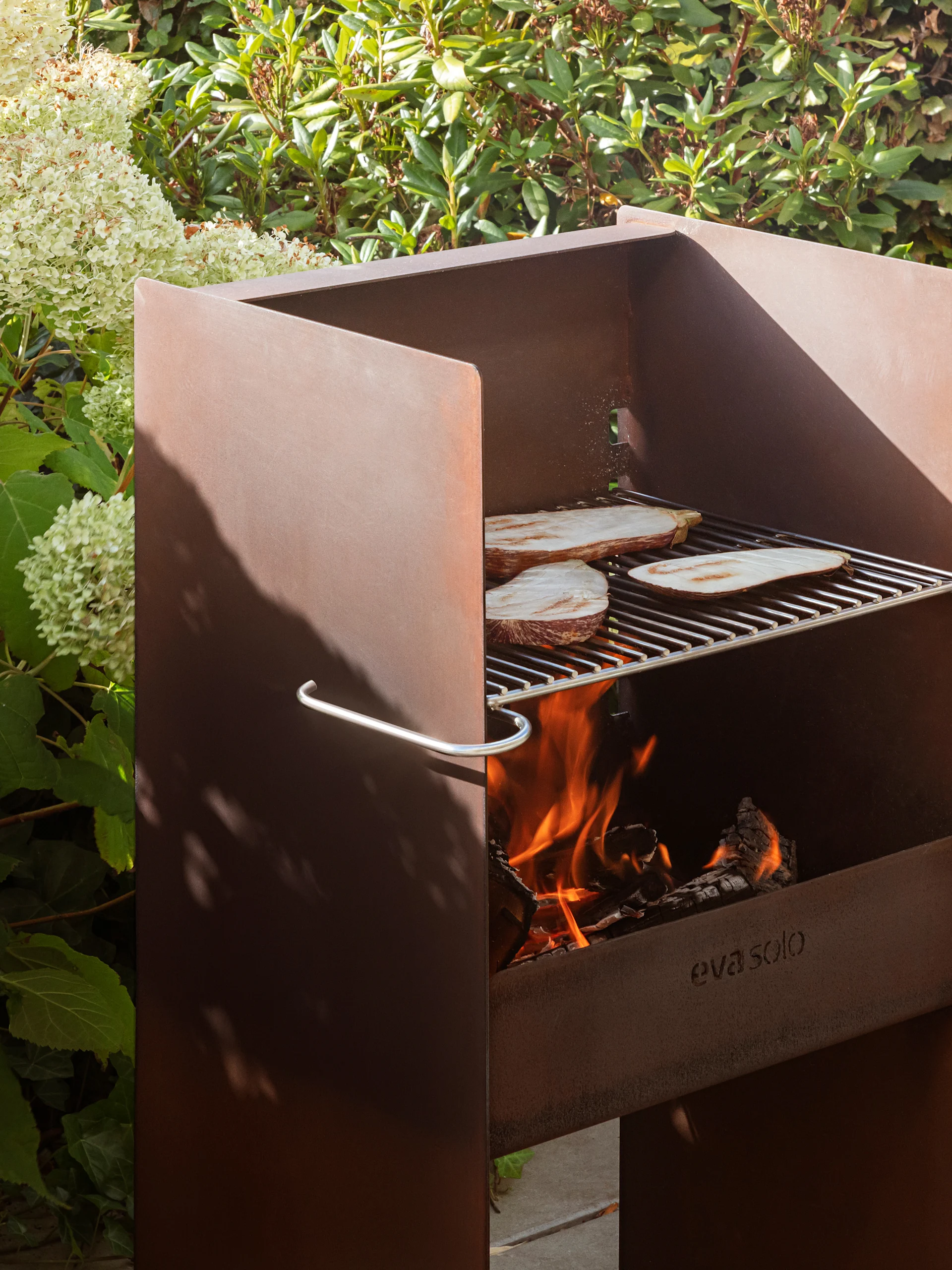 Verroeste metalen buitenbarbecue van Eva Solo met aubergine op het rooster, brandend vuur en weelderig groen.