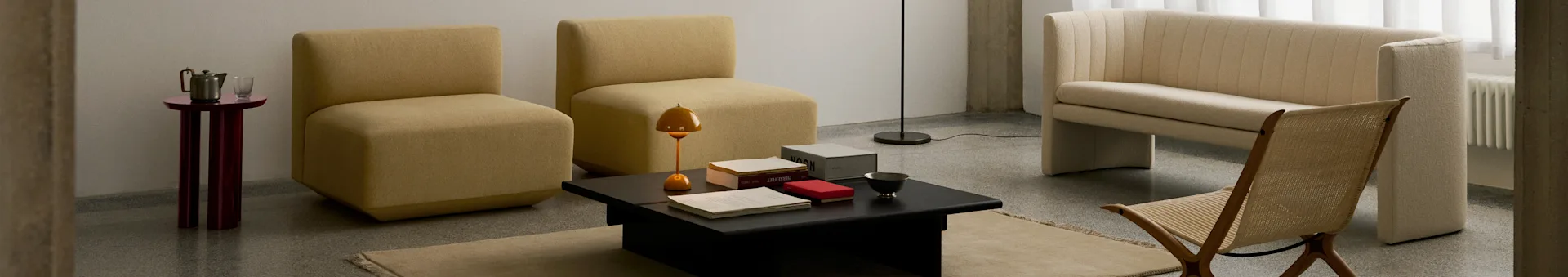 Een moderne woonkamer met twee mosterdkleurige armloze stoelen, een zwarte salontafel, een crèmekleurige geribbelde bank en een gevlochten houten stoel.