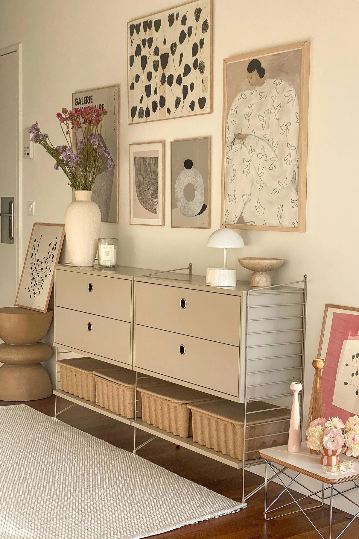 Een moderne kamer met een beige modulair dressoir, laden en opbergmanden. Een vaas met bloemen, lamp en abstracte kunst sieren de ruimte.