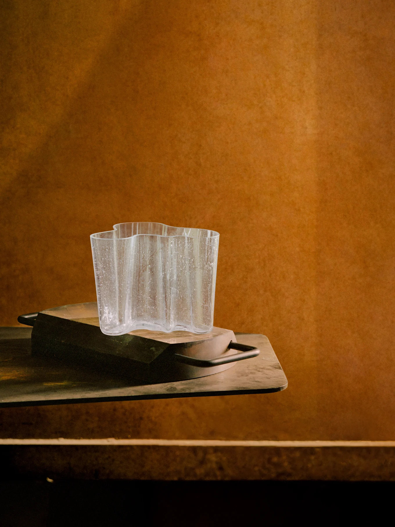 Heldere, golvende glazen vaas met waterdruppels op een donker houten dienblad, tegen een warme bruine getextureerde achtergrond.