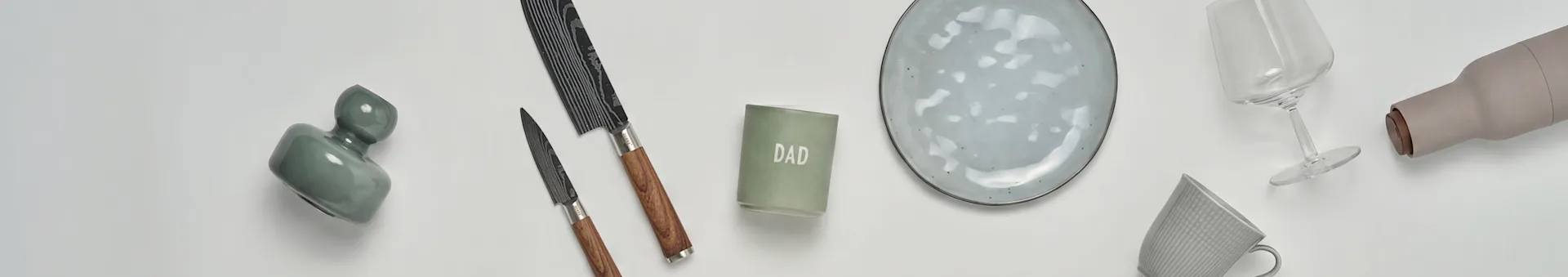 Een flat lay van keuken- en cadeauartikelen: een groene vaas, twee messen, een 'DAD'-kaars, een grijs keramisch bord, een wijnglas, een beige fles en een grijze mok.